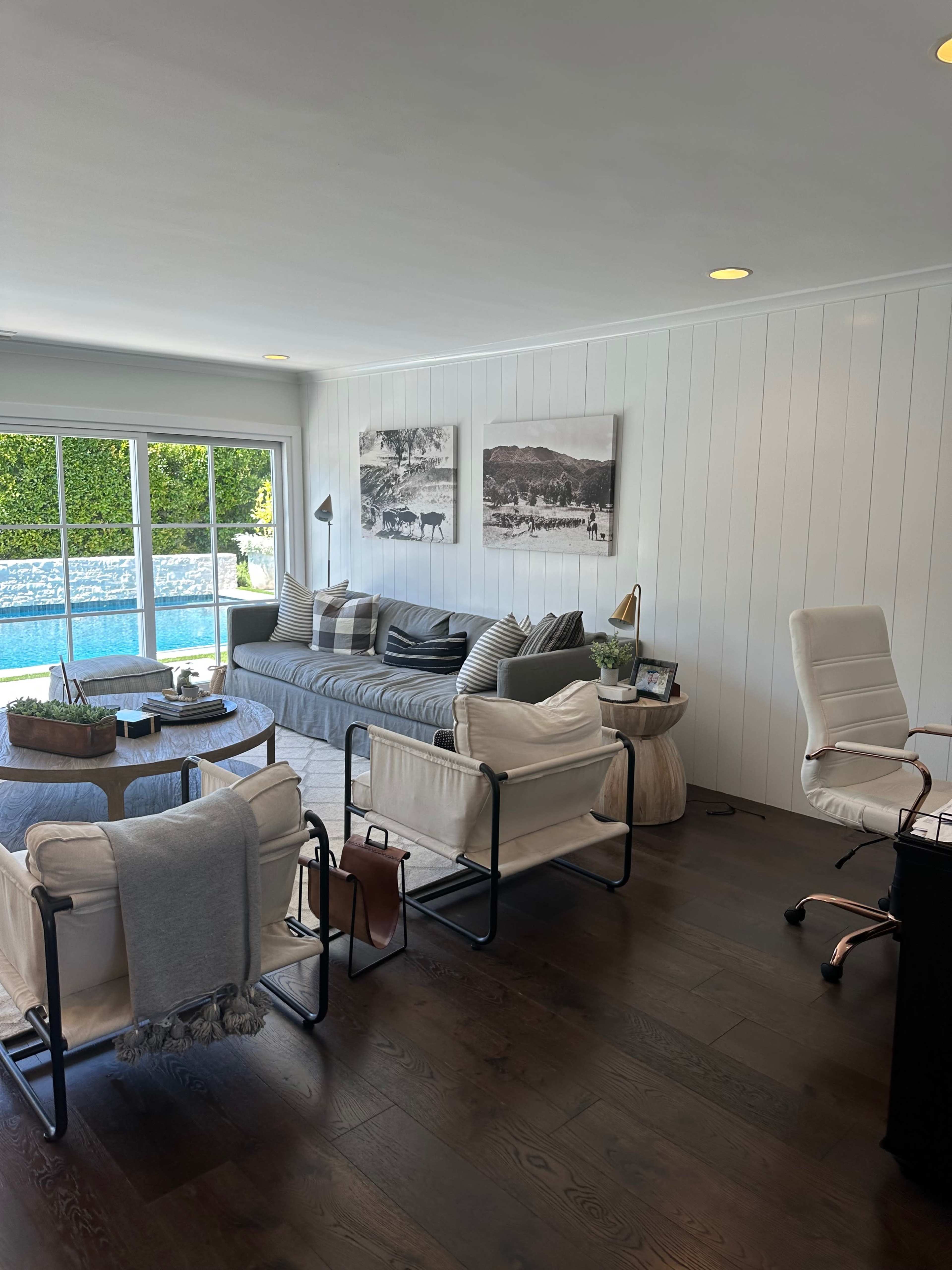 A modern living room features a gray sofa, two white chairs, and large windows overlooking a pool area, with artwork hanging on the walls.