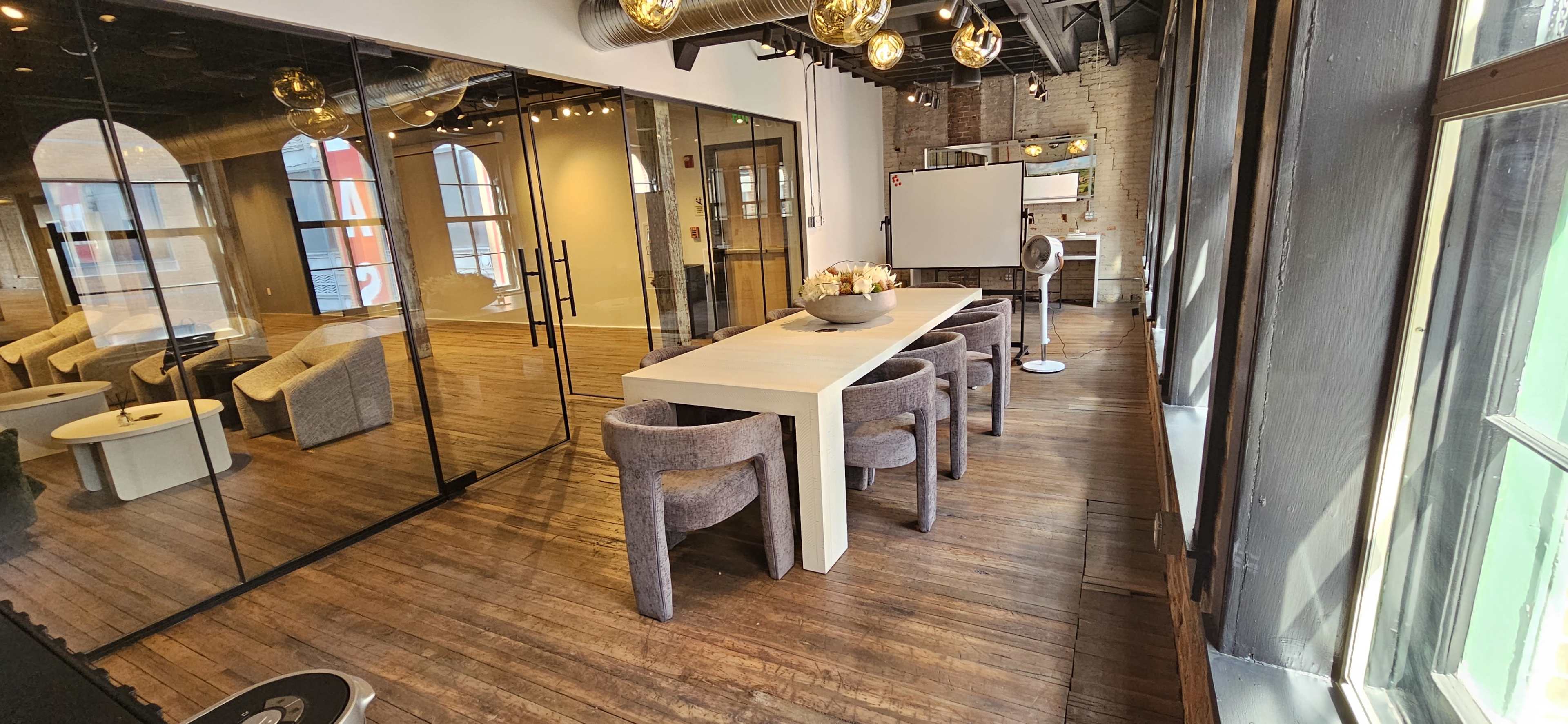 A modern office space featuring a long white table surrounded by gray chairs, with large glass walls and wooden flooring.
