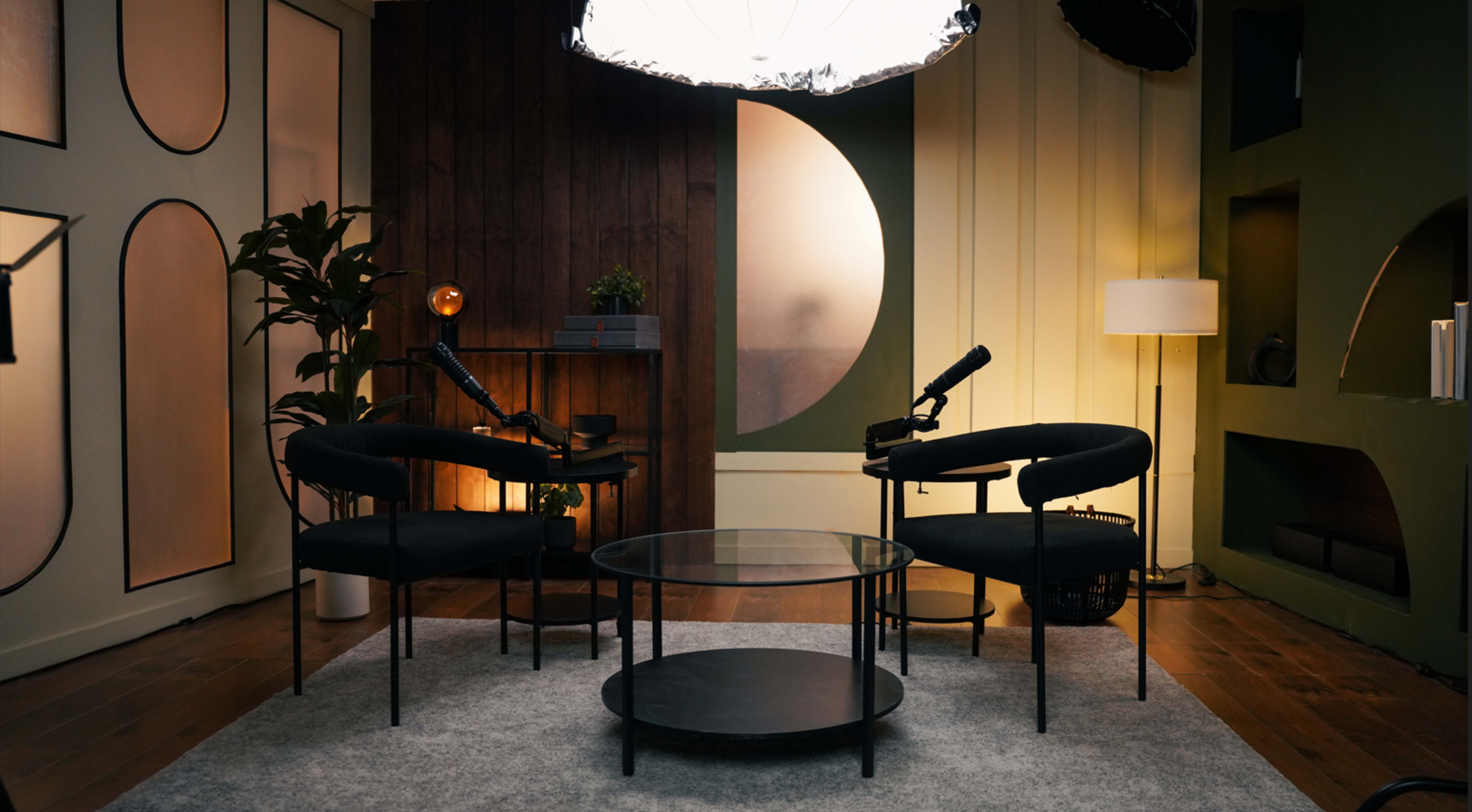 The image shows a cozy, modern interior with two black chairs facing a small glass table, under a large, circular light fixture.