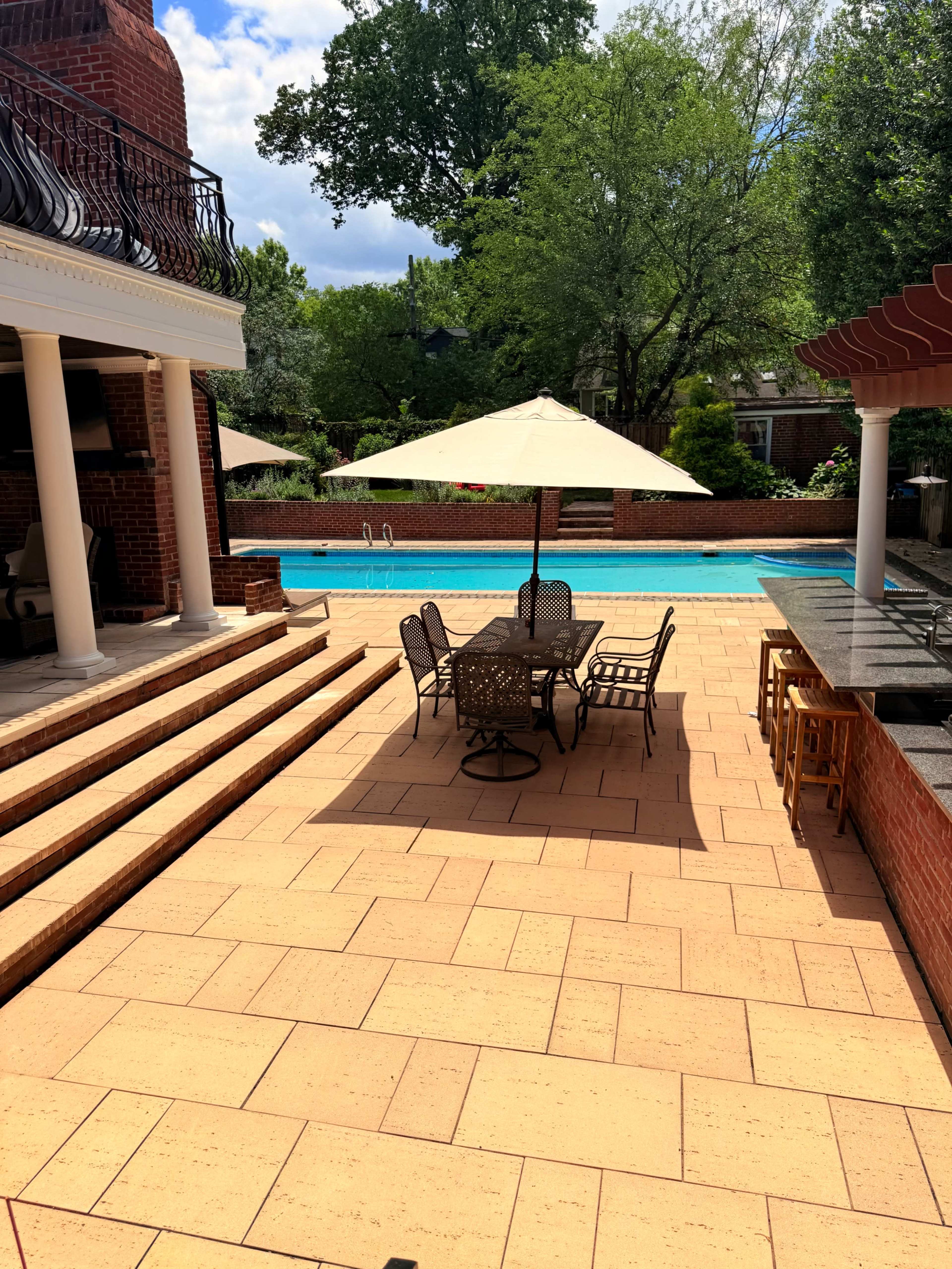 The image shows a spacious outdoor patio with a swimming pool, dining set, and several umbrellas surrounded by greenery and a brick house.