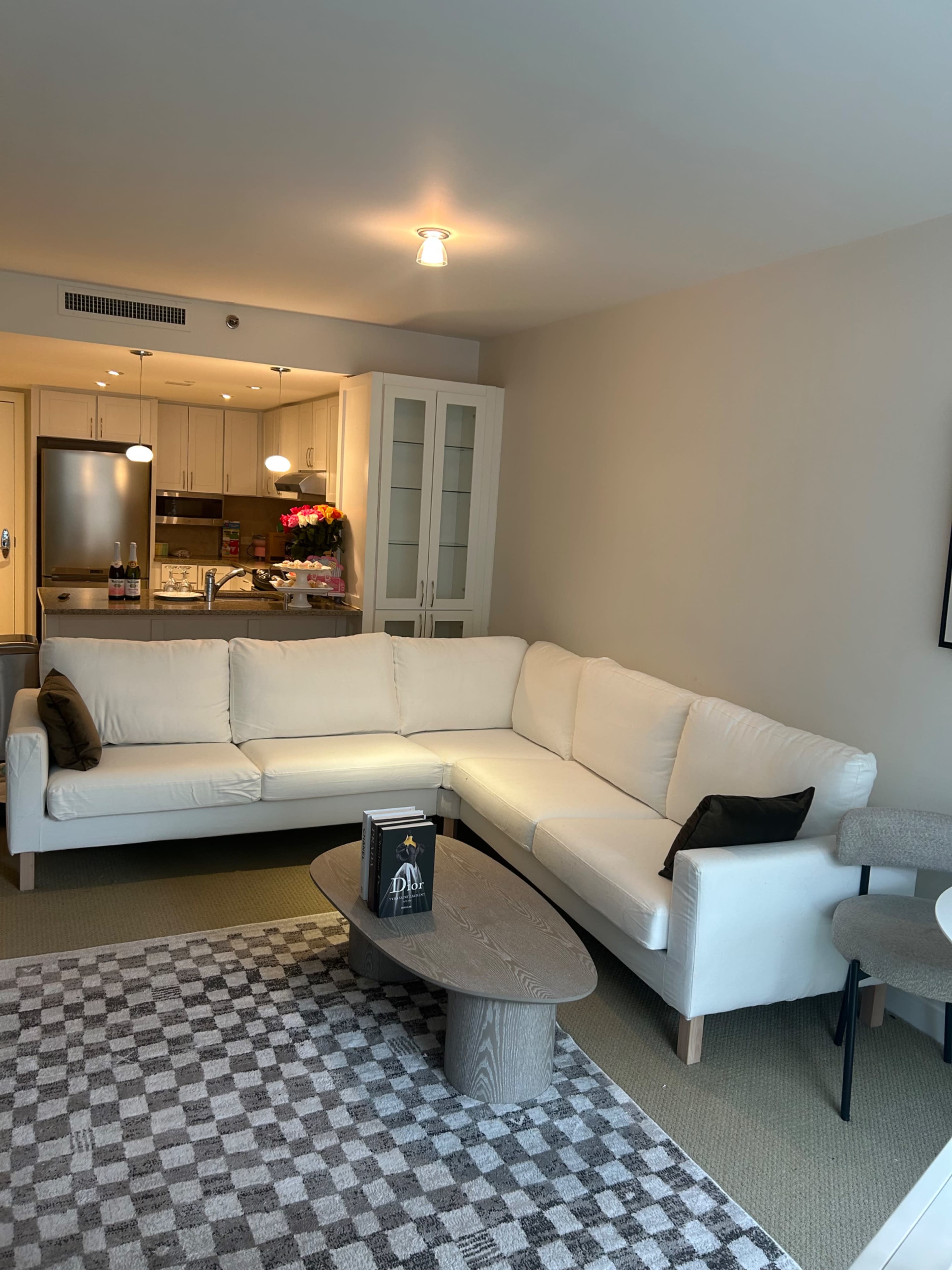 The image shows a modern living room featuring a white sectional sofa, a round coffee table, and a neatly arranged area rug, with a kitchen visible in the background.