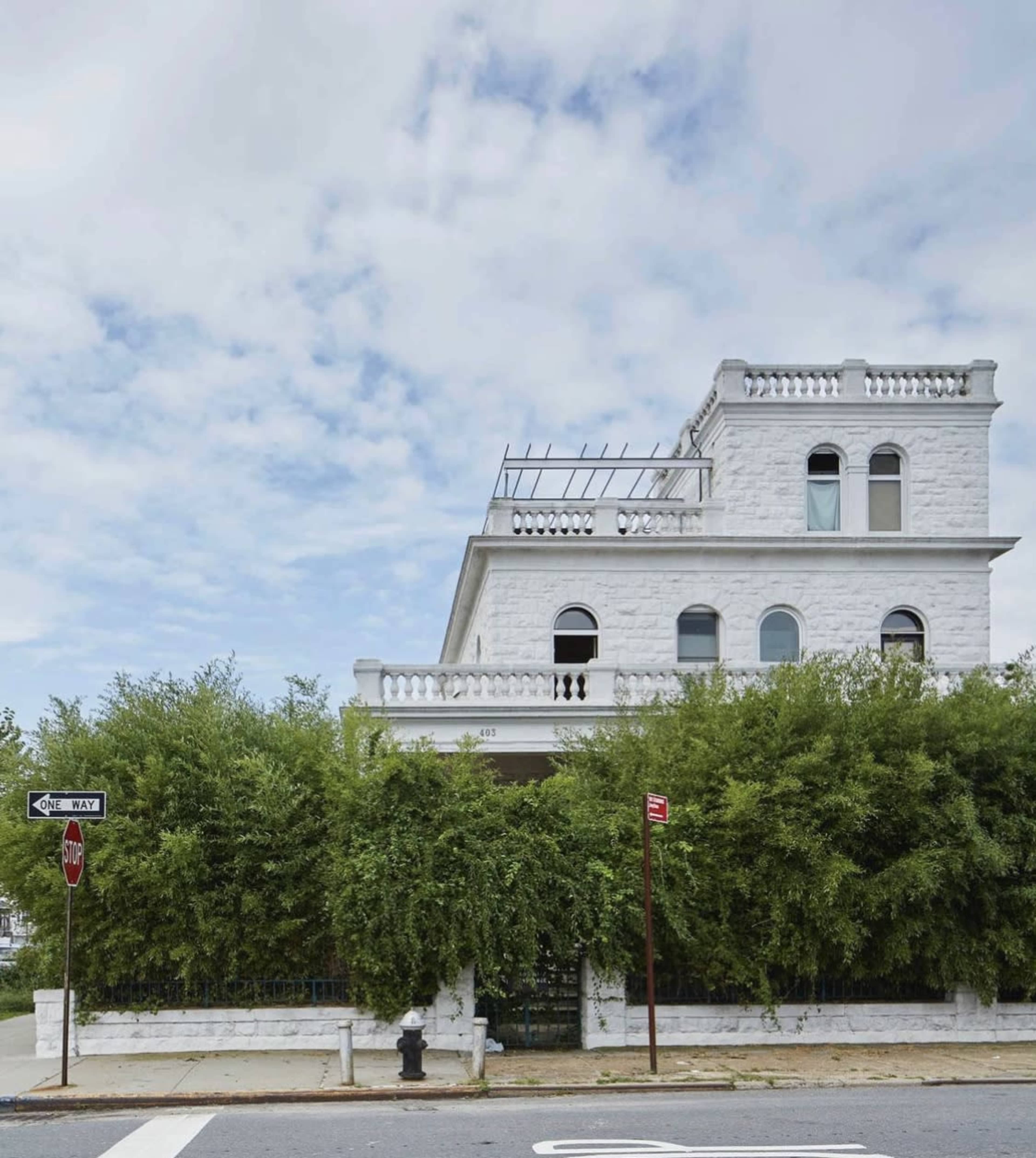 A large white building with multiple windows stands behind a tall green hedge, located at the corner of a street marked by a stop sign and a no parking sign.