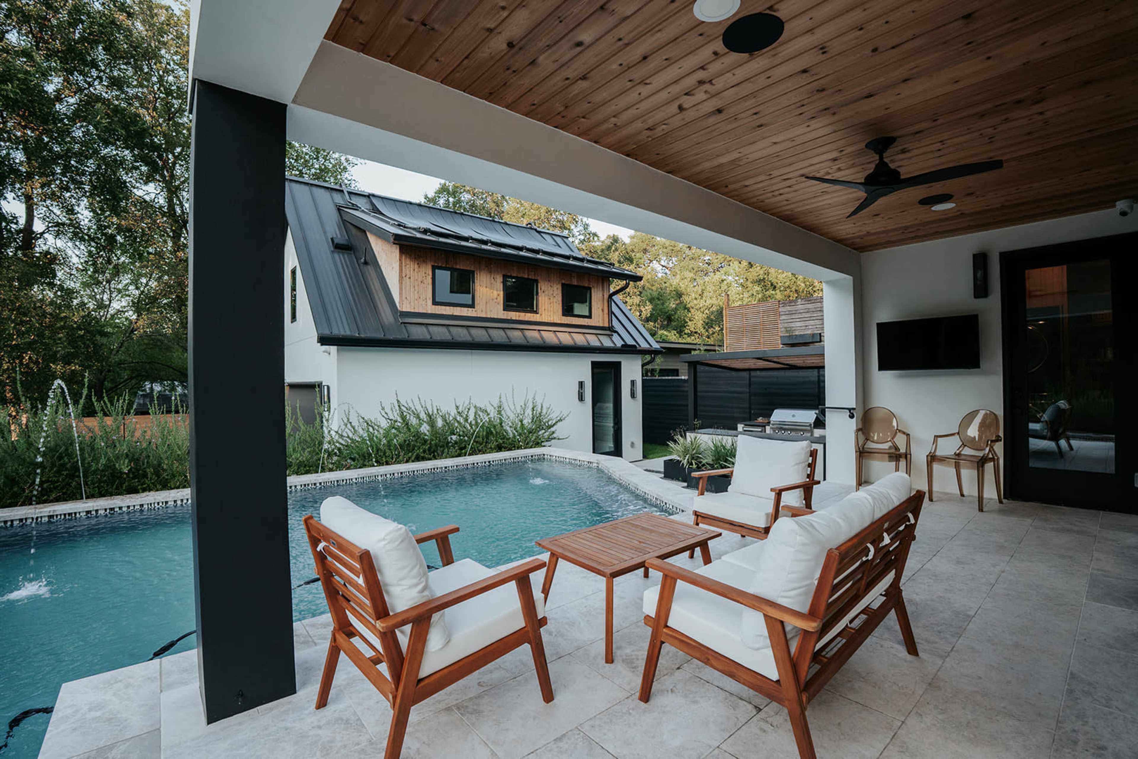 The image shows a modern outdoor living space featuring a swimming pool, wooden lounge chairs, and a small wooden table, with a contemporary house in the background.