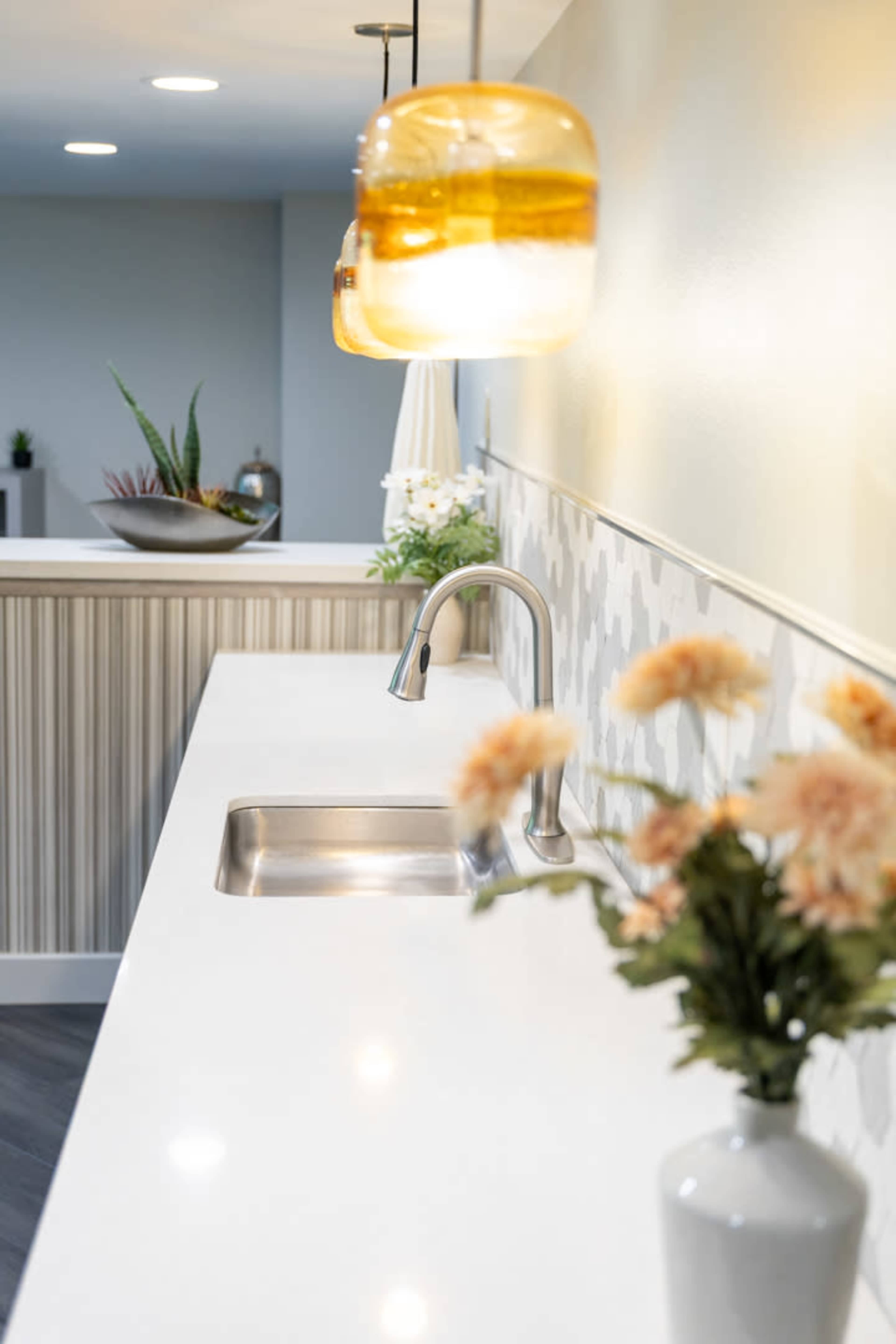 The image shows a modern kitchen counter with a stainless steel sink, a flower vase, and decorative pendant lights above.