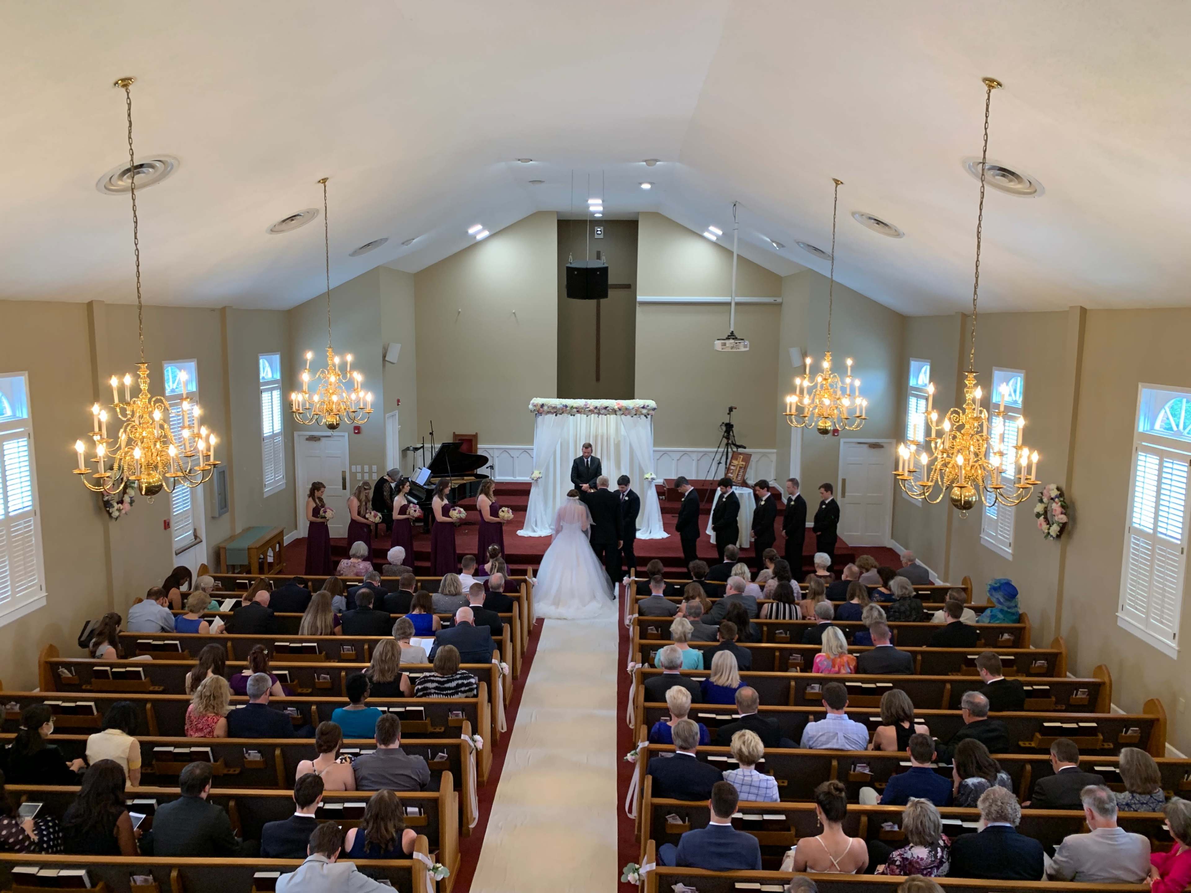 A wedding ceremony is taking place in a church filled with seated guests, while the bridal couple stands at the altar surrounded by attendants.