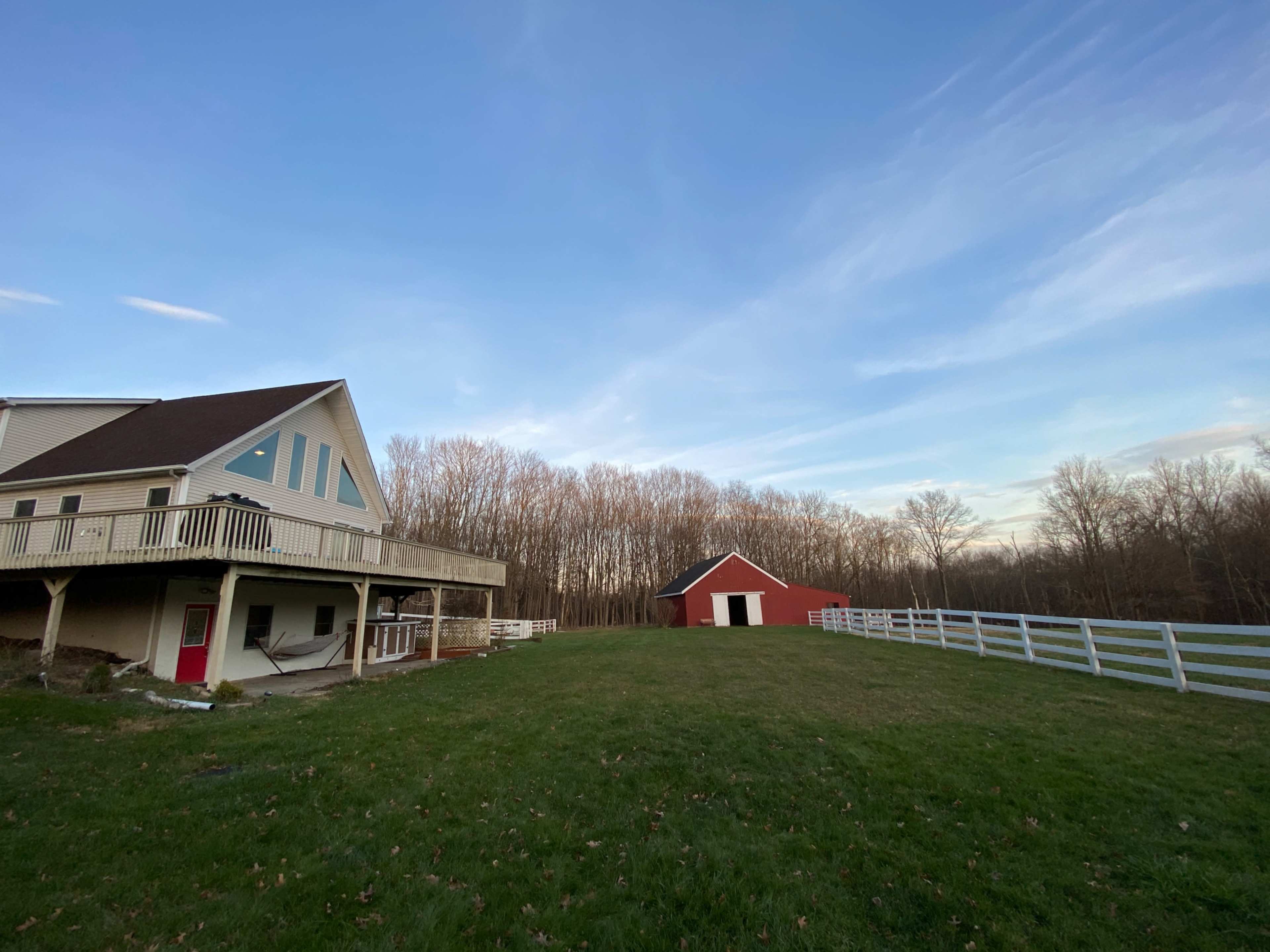 A two-story house with a deck stands next to a red barn in a grassy field surrounded by trees.