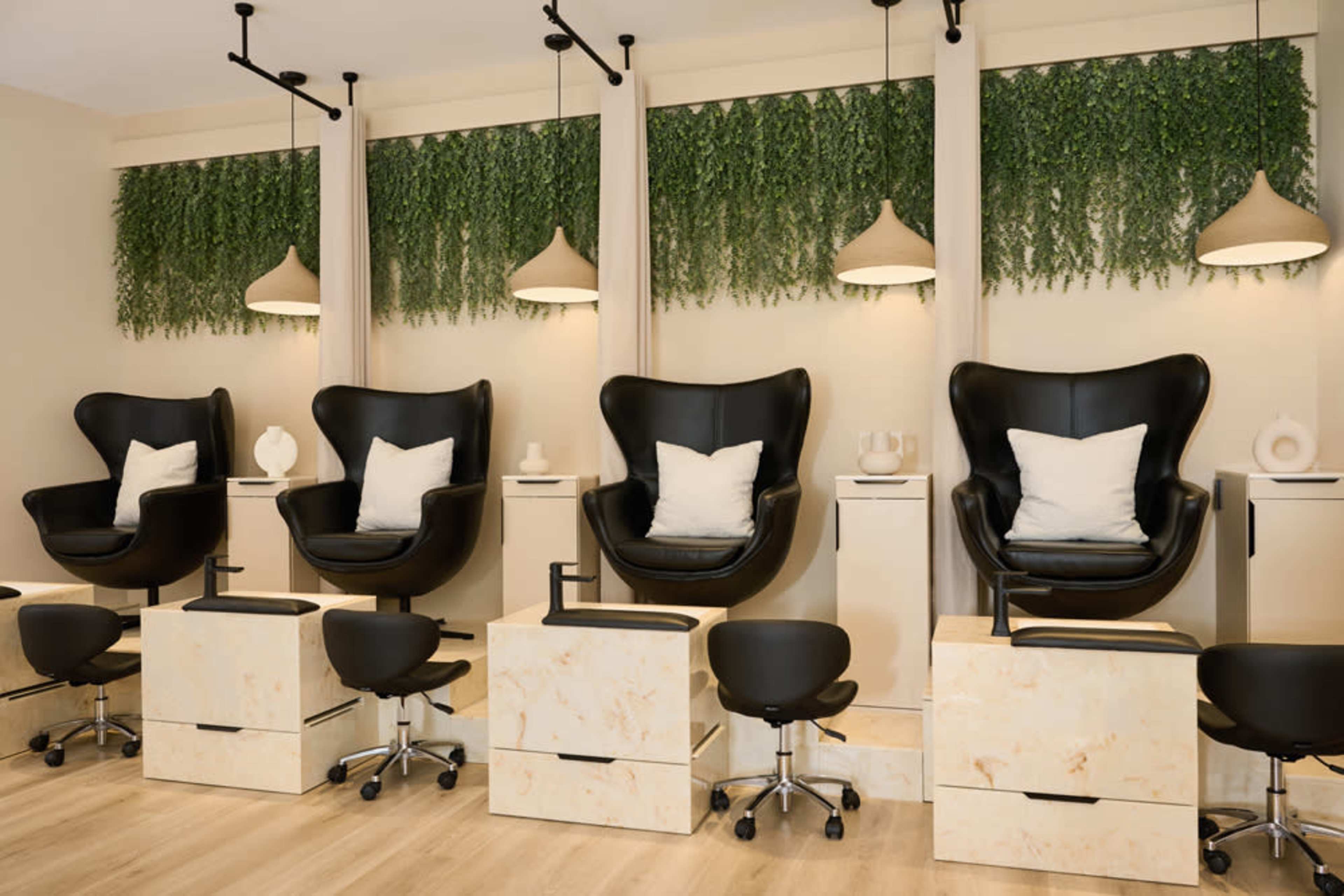 The image shows a nail salon interior featuring four black salon chairs on platforms, each accompanied by a small rolling stool, with greenery and modern light fixtures above.