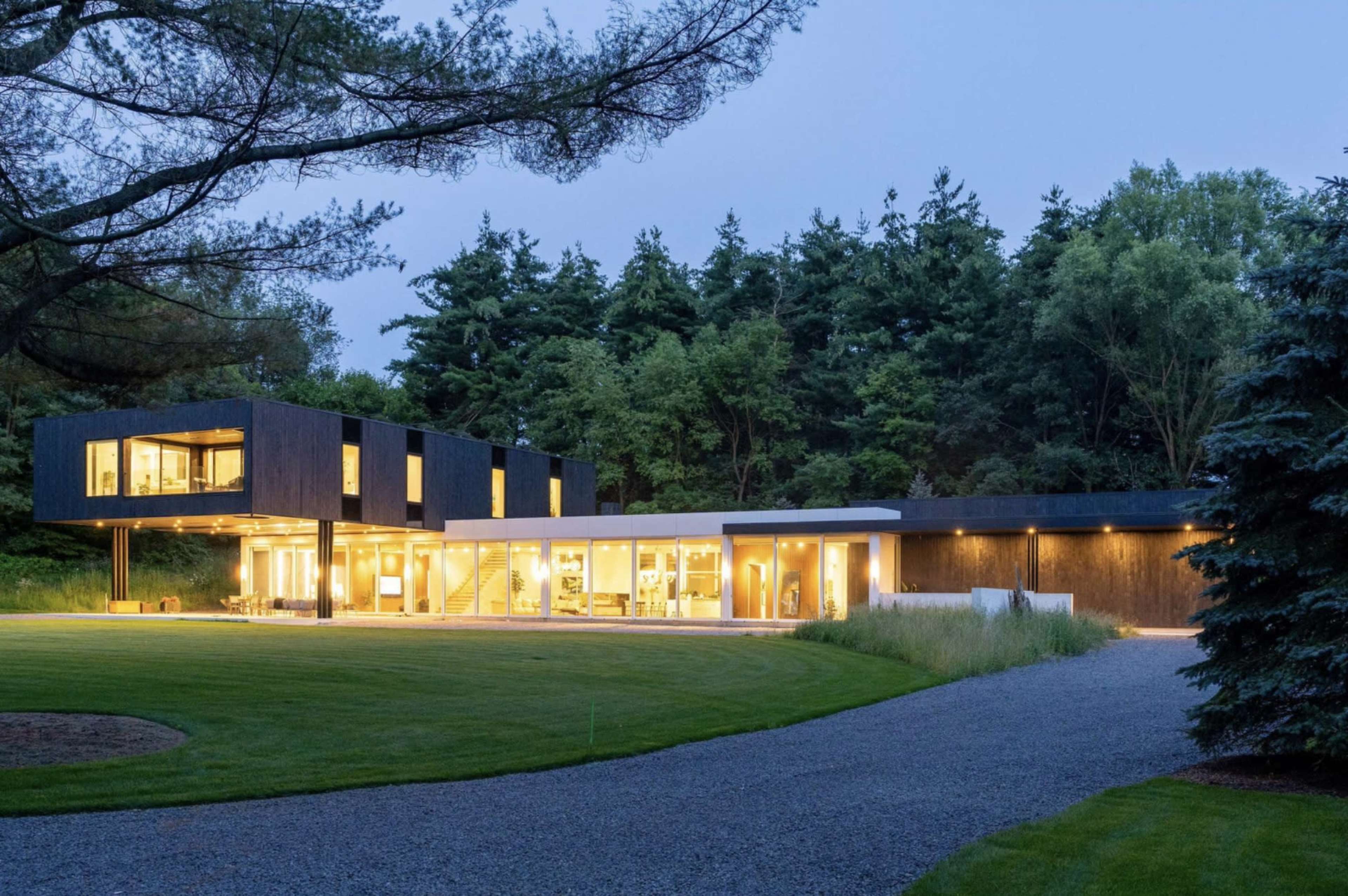 A modern two-story house with large windows, situated in a landscaped area with trees and a gravel driveway.