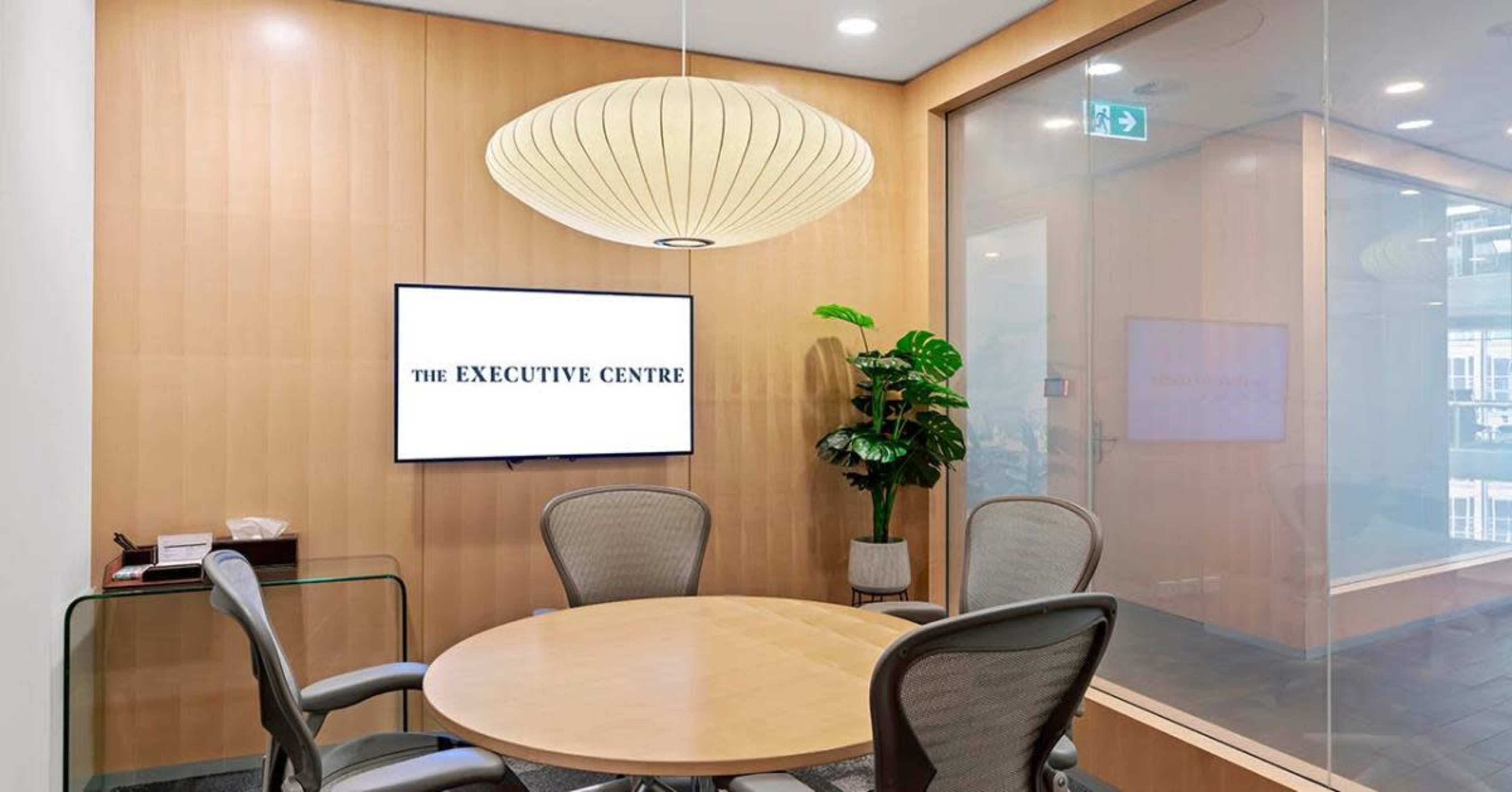 A minimalist meeting room features a round wooden table, ergonomic chairs, a wall-mounted television displaying "THE EXECUTIVE CENTRE," and a large pendant light above.