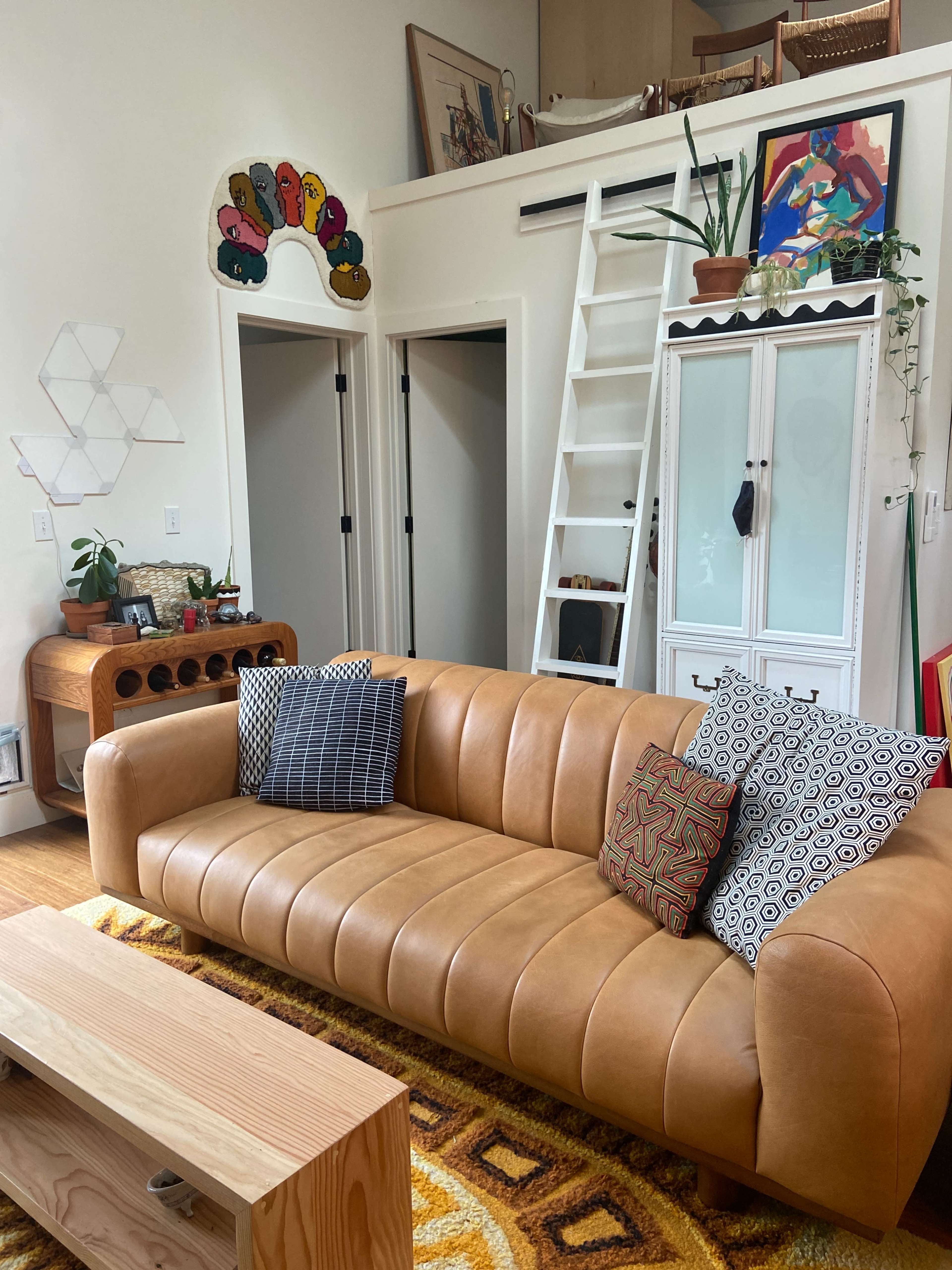 The scene features a modern living room with a tan leather sofa, a wooden coffee table, and colorful wall art above a staircase leading to a loft area.