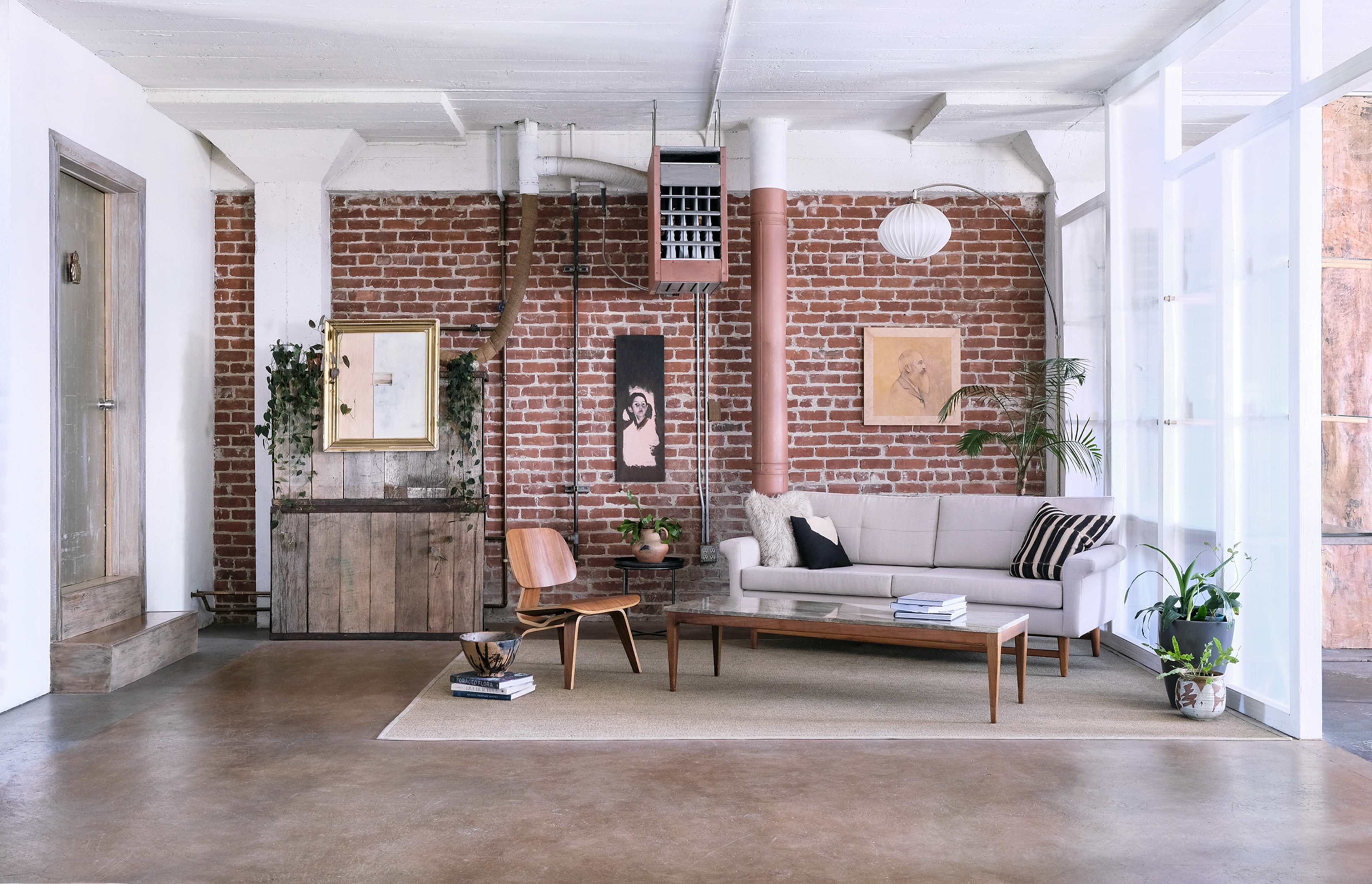 A minimalistic living room with a brick wall, a light-colored couch, a wooden coffee table, and various decorative plants and artwork.