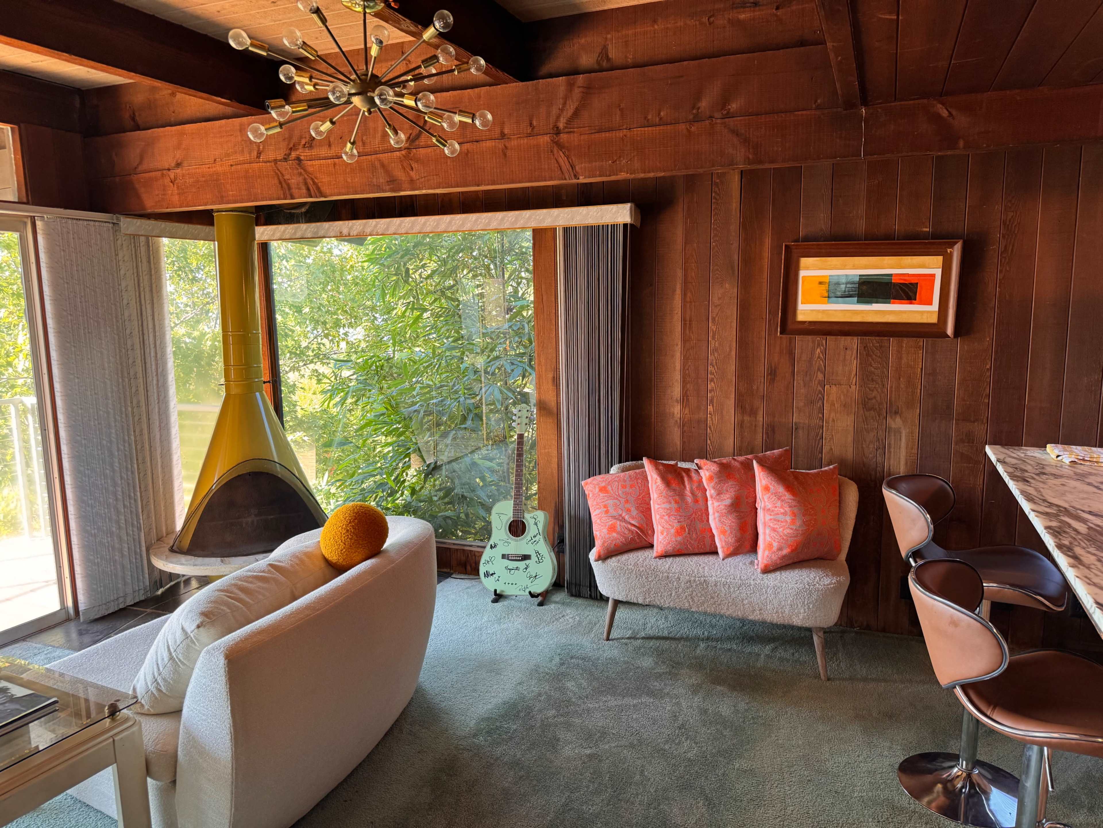 A cozy living room features a circular pendant light, a yellow fireplace, a gray sofa with orange pillows, and a view of greenery through large windows.