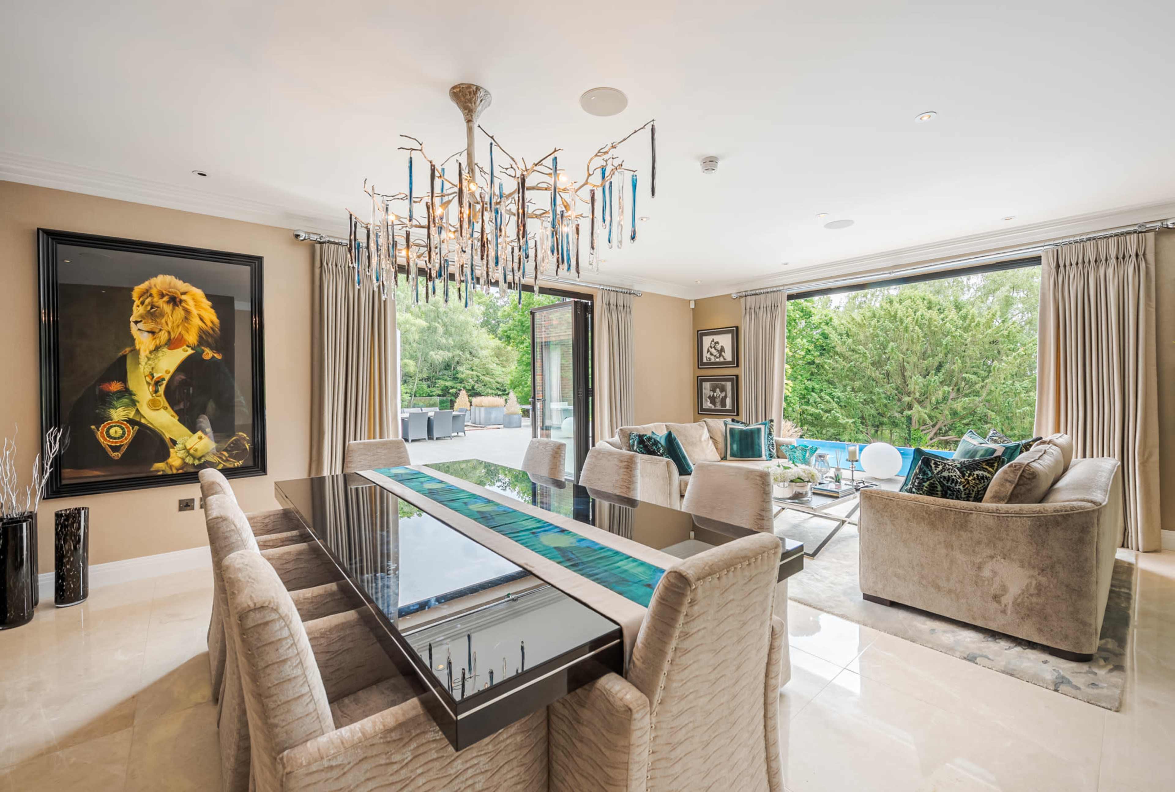 A modern dining room features a long glass table surrounded by upholstered chairs, with a large artwork of a lion on the wall and large windows overlooking a garden.