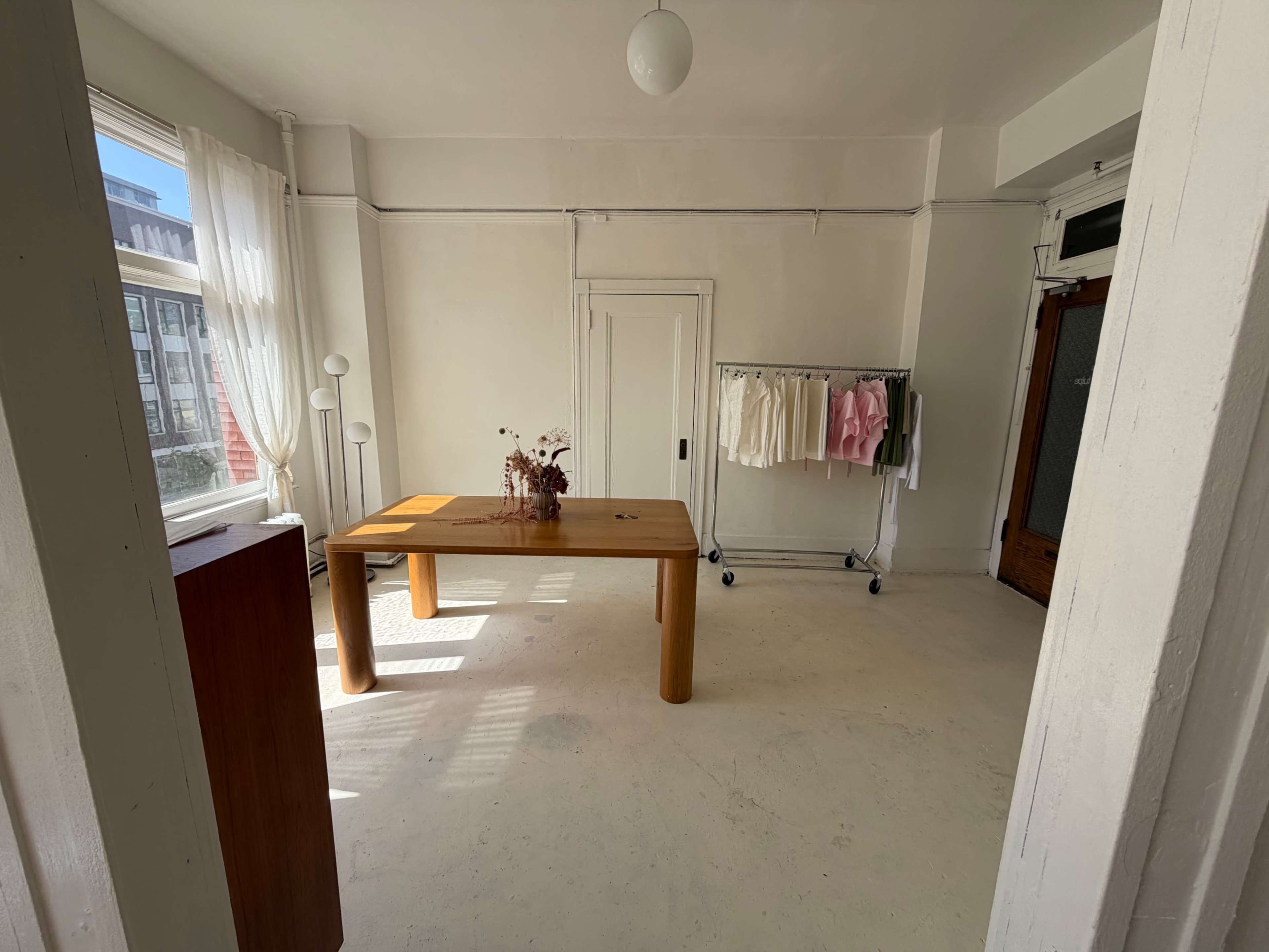 The room features a wooden table in the center, surrounded by light-colored walls, a clothing rack with garments, and a window allowing natural light to enter.