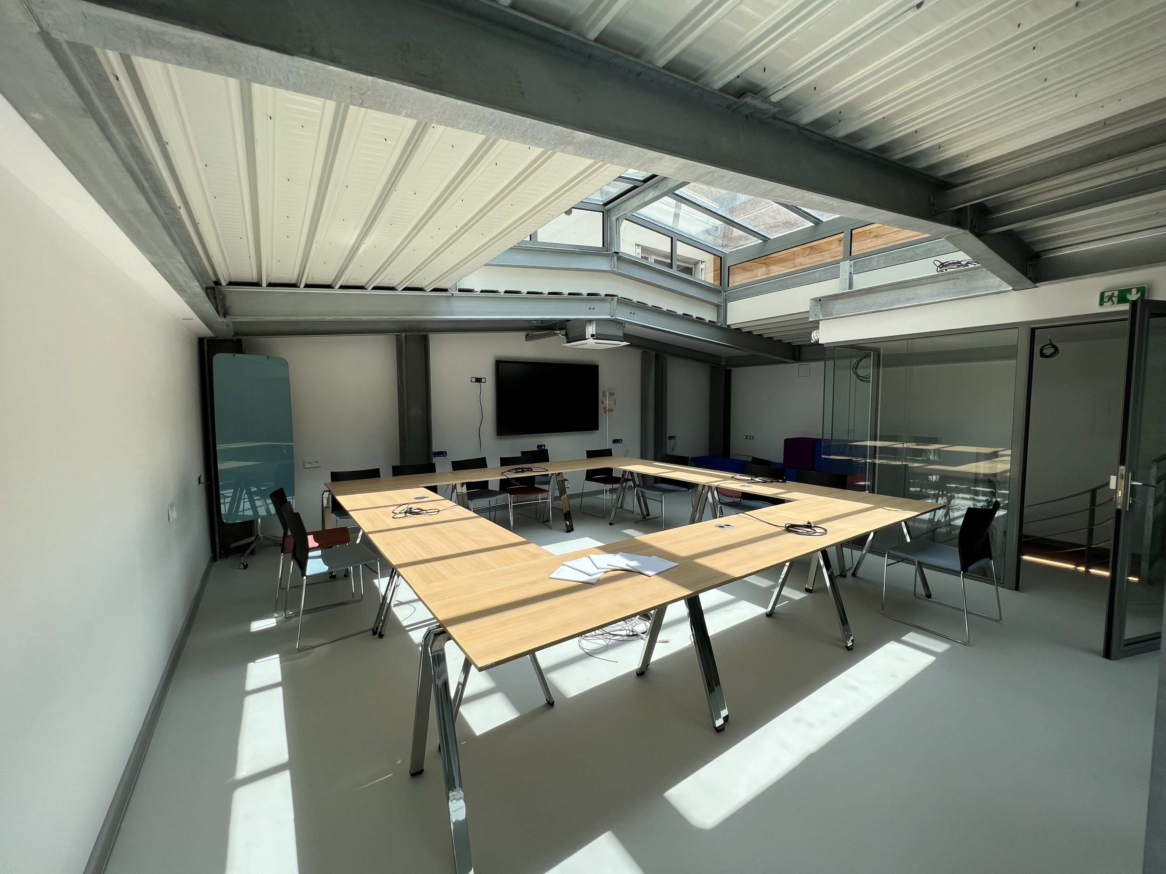 A bright meeting room features a large wooden table surrounded by chairs, with a television on the wall and natural light coming through a skylight.