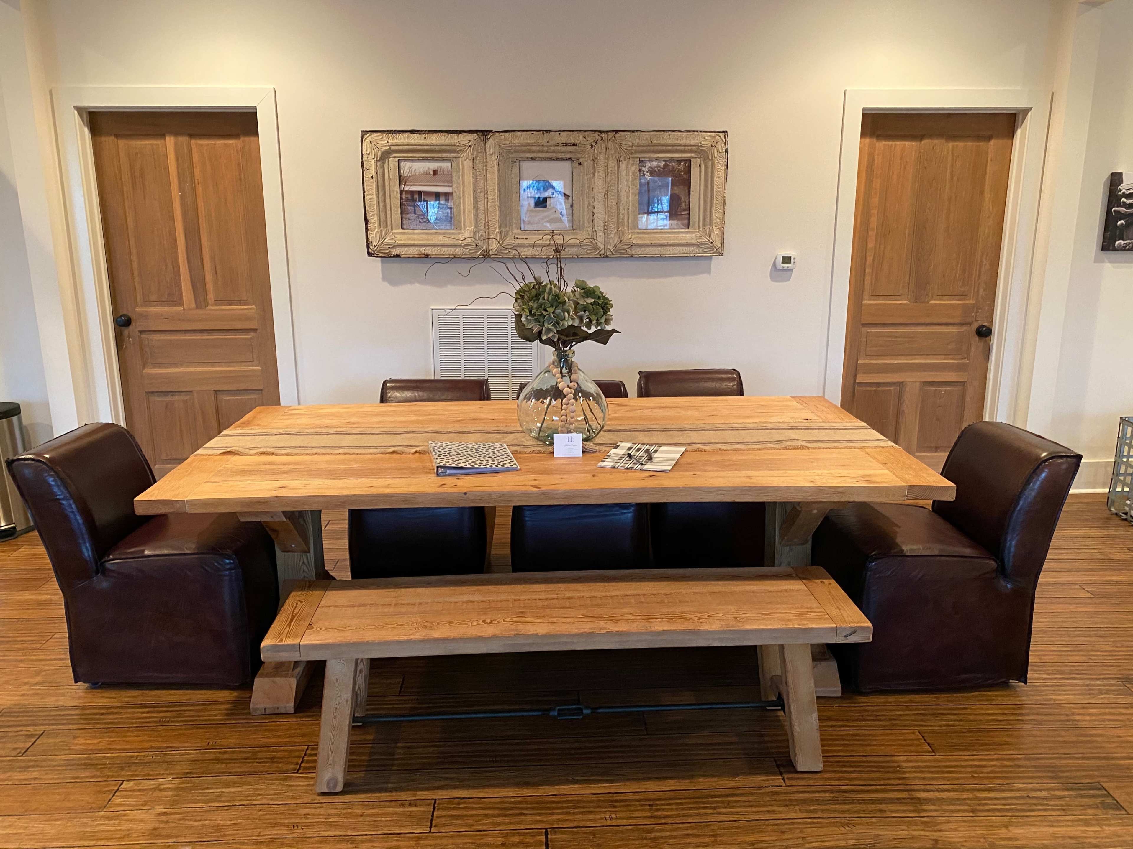 A wooden dining table with a vase and magazines is surrounded by two brown leather chairs and a bench in a room with two wooden doors.