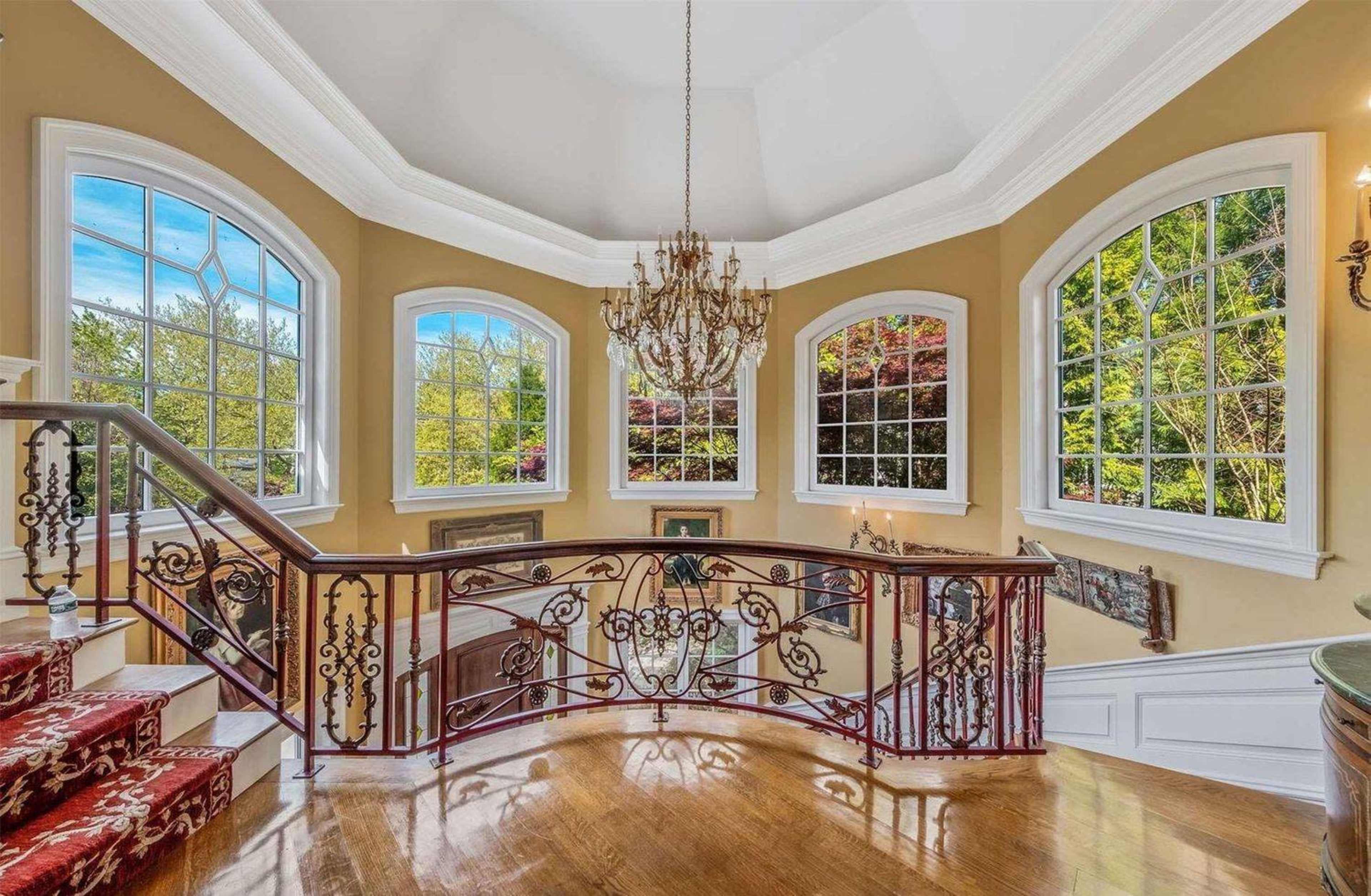 The image shows a well-lit foyer featuring a curved staircase with an ornate railing, large windows allowing natural light, and a chandelier overhead.