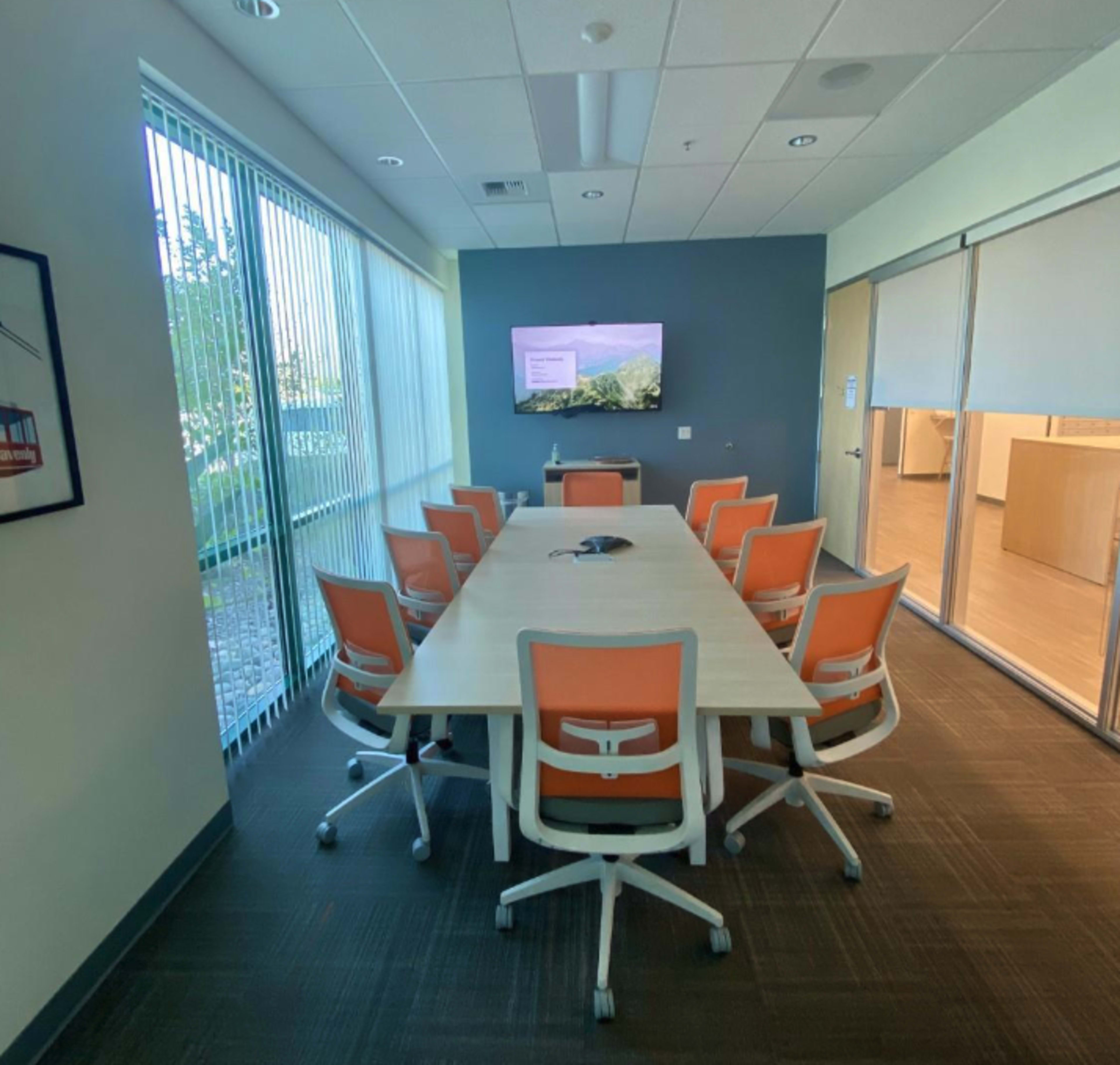 A conference room featuring a long table surrounded by eight orange and white chairs, with a wall-mounted screen displaying a nature image and large windows providing natural light.