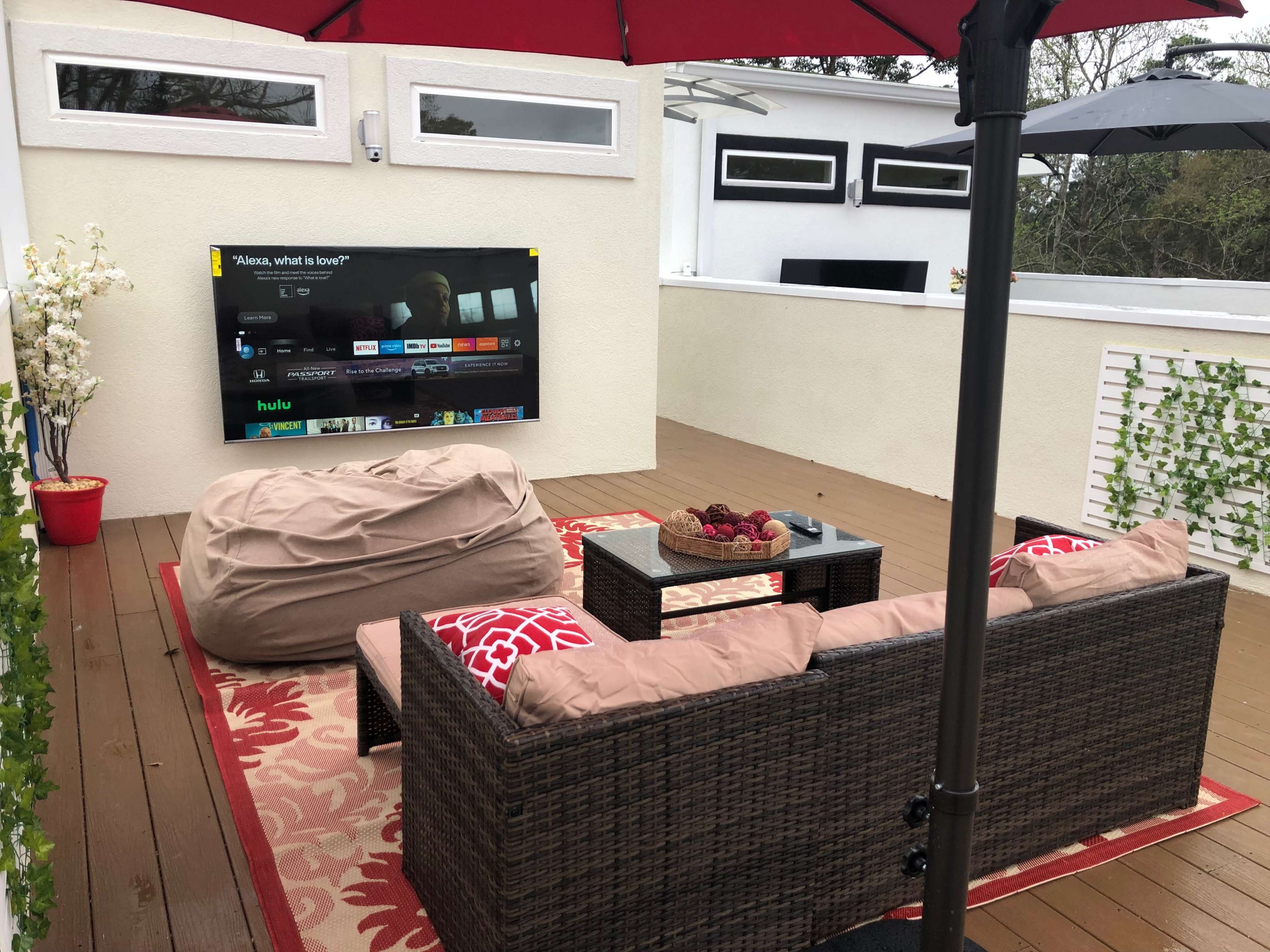 The outdoor seating area features a television mounted on a wall, surrounded by a coffee table, bean bags, and a couch, all beneath a red umbrella.