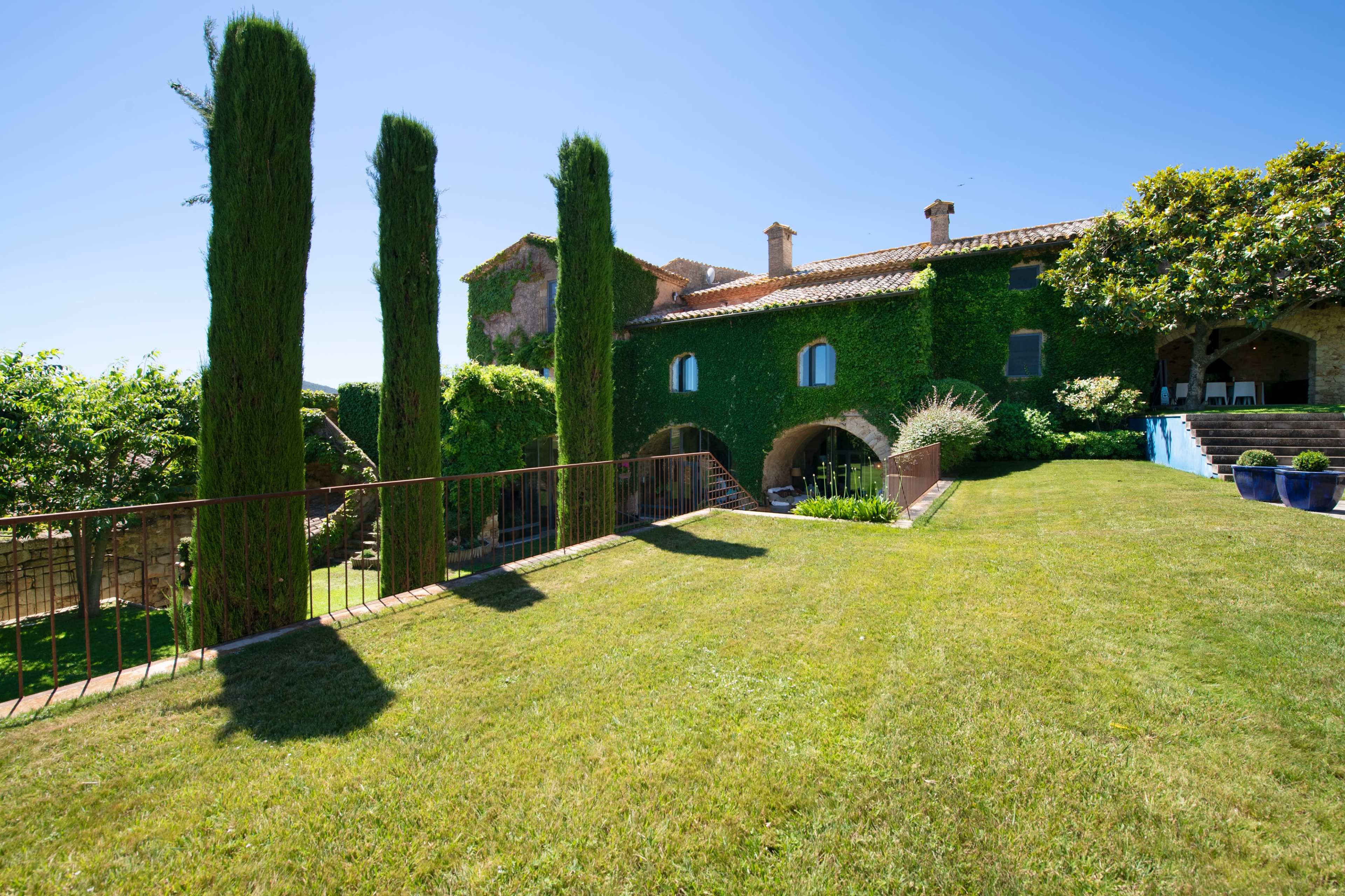 A stone house covered in ivy stands beside tall cypress trees on a well-maintained lawn under a clear blue sky.
