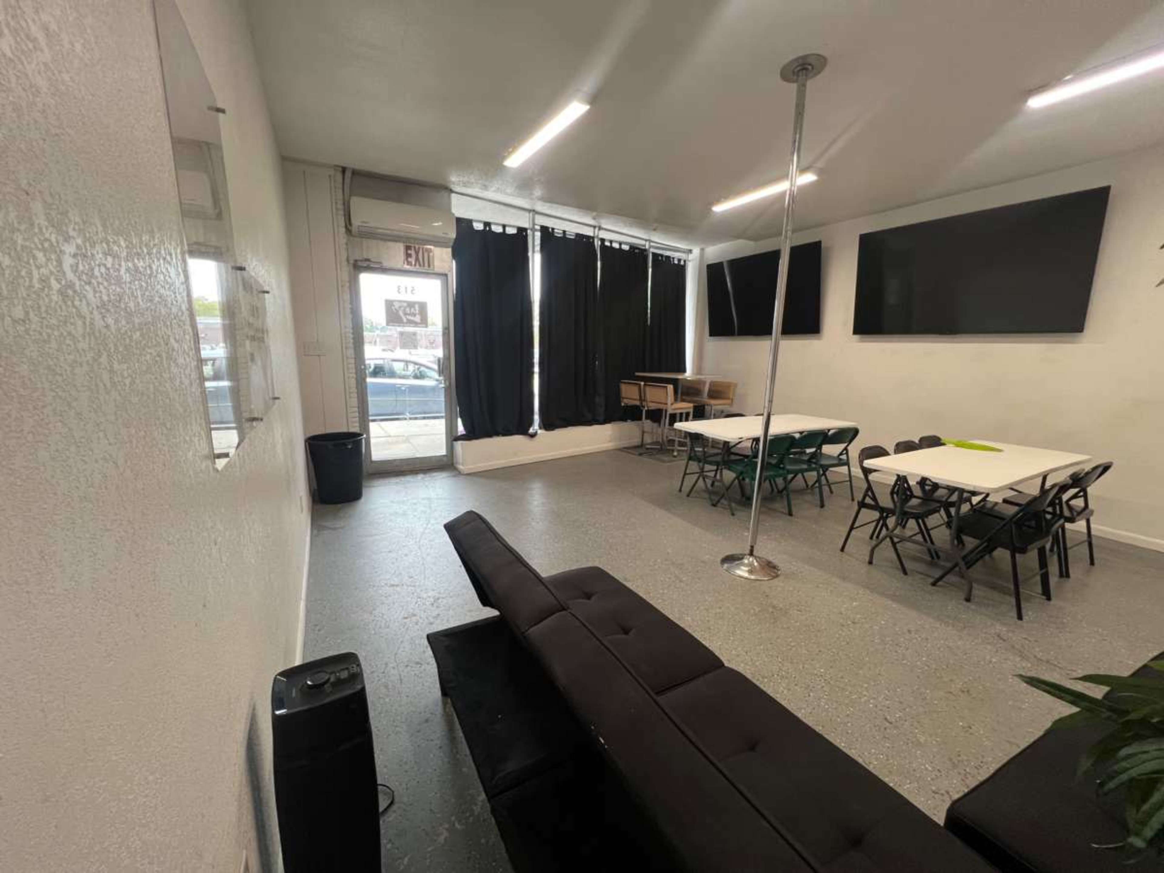 The image shows a spacious room with a black sofa, tables and chairs arranged for seating, and large screens mounted on the walls.