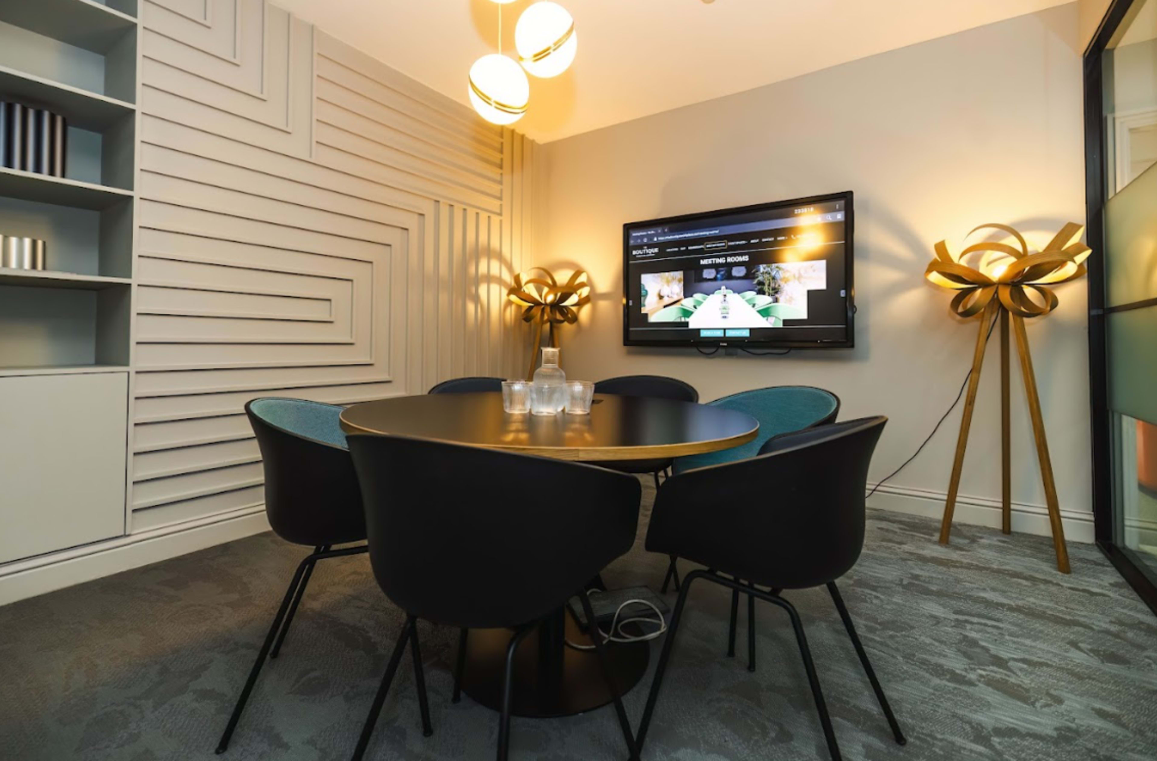 The image shows a modern meeting room with a circular table surrounded by black chairs, a television mounted on the wall, and contemporary light fixtures.