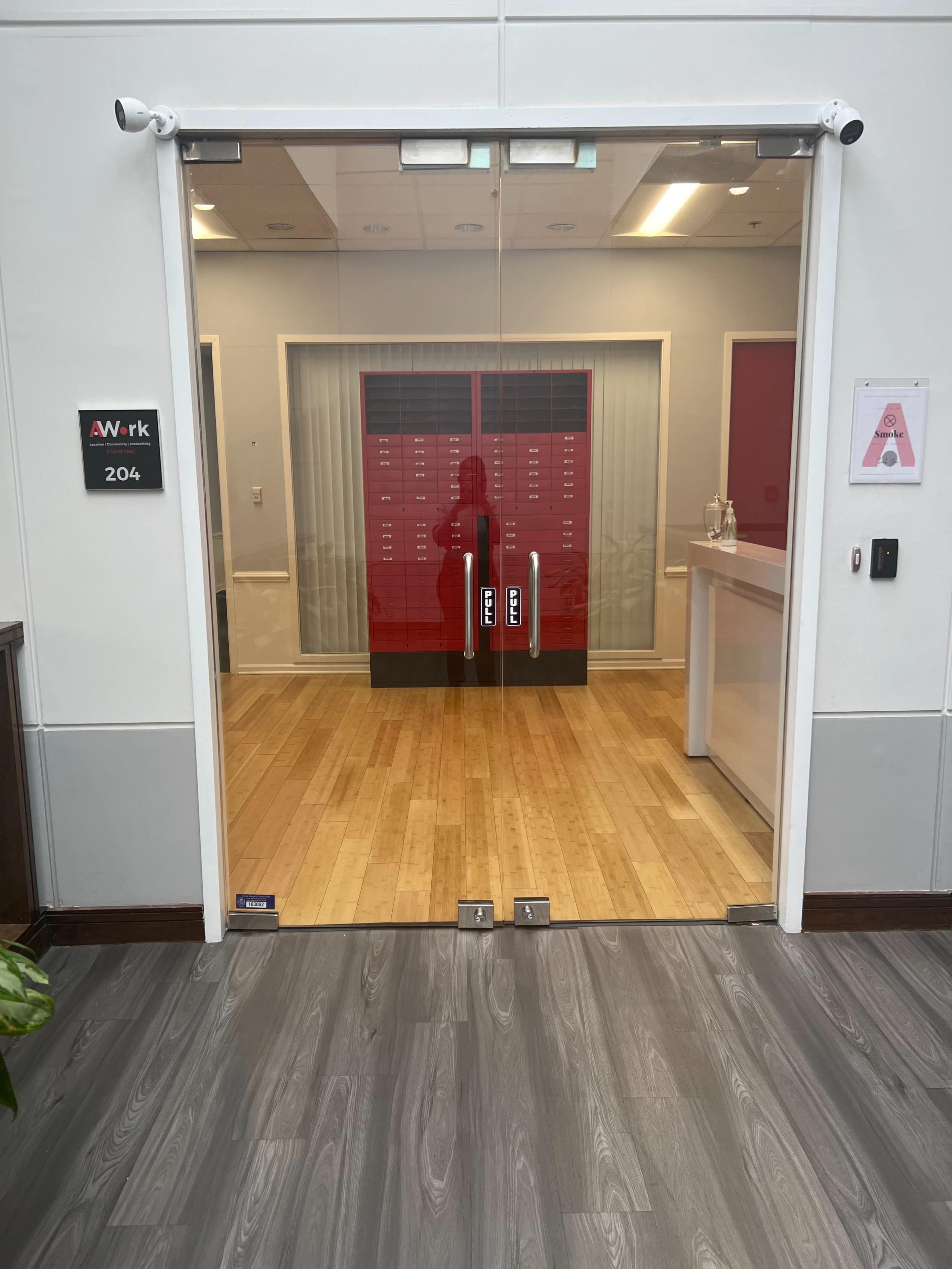 A glass door with a sign labeled "204" leading to a brightly lit office area featuring red lockers and wooden flooring.