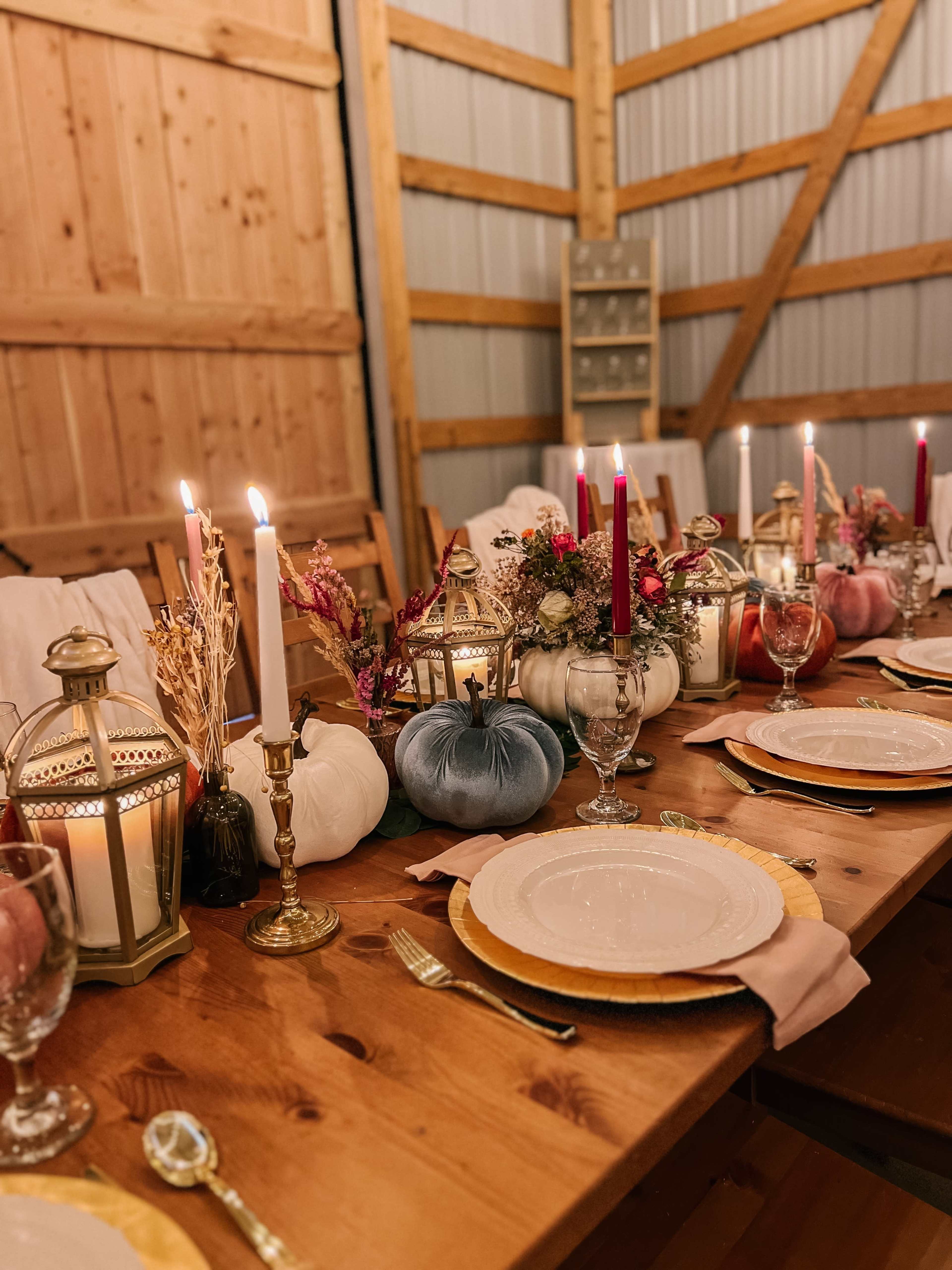 A rustic wooden dining table is set with white plates, gold chargers, and decorative pumpkins, flanked by candle holders and floral arrangements.