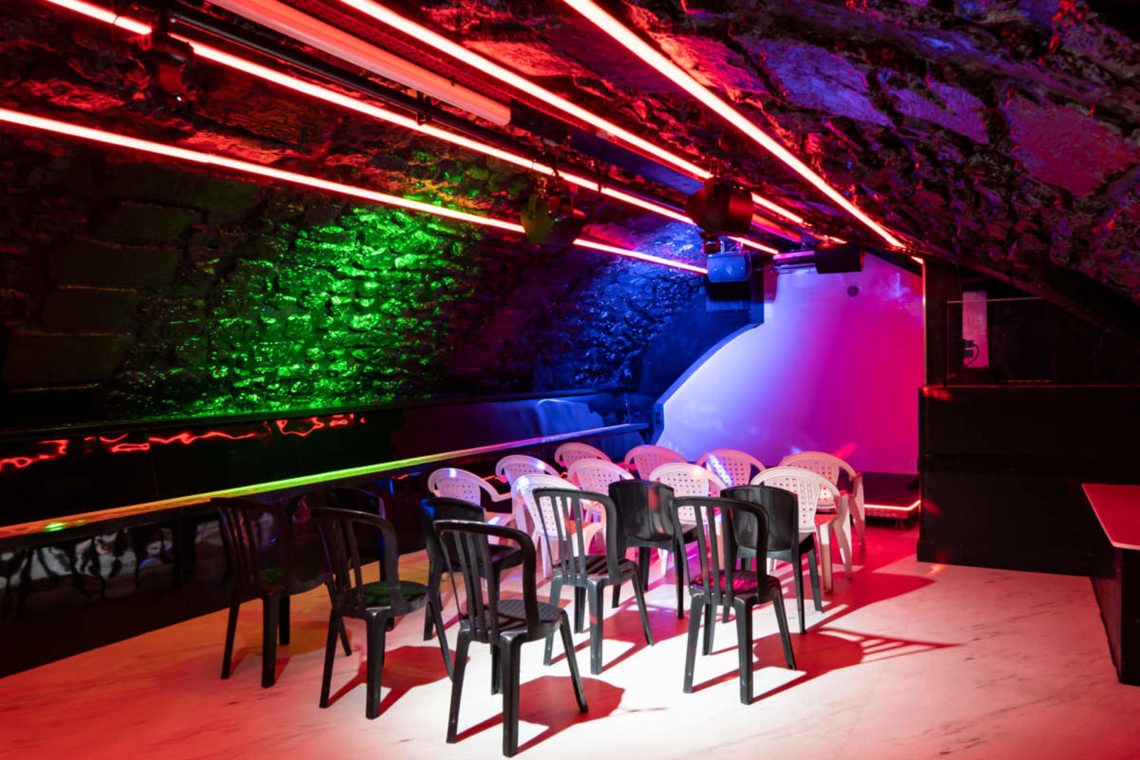 A dimly lit underground venue features a row of white and black chairs under vibrant neon lighting.