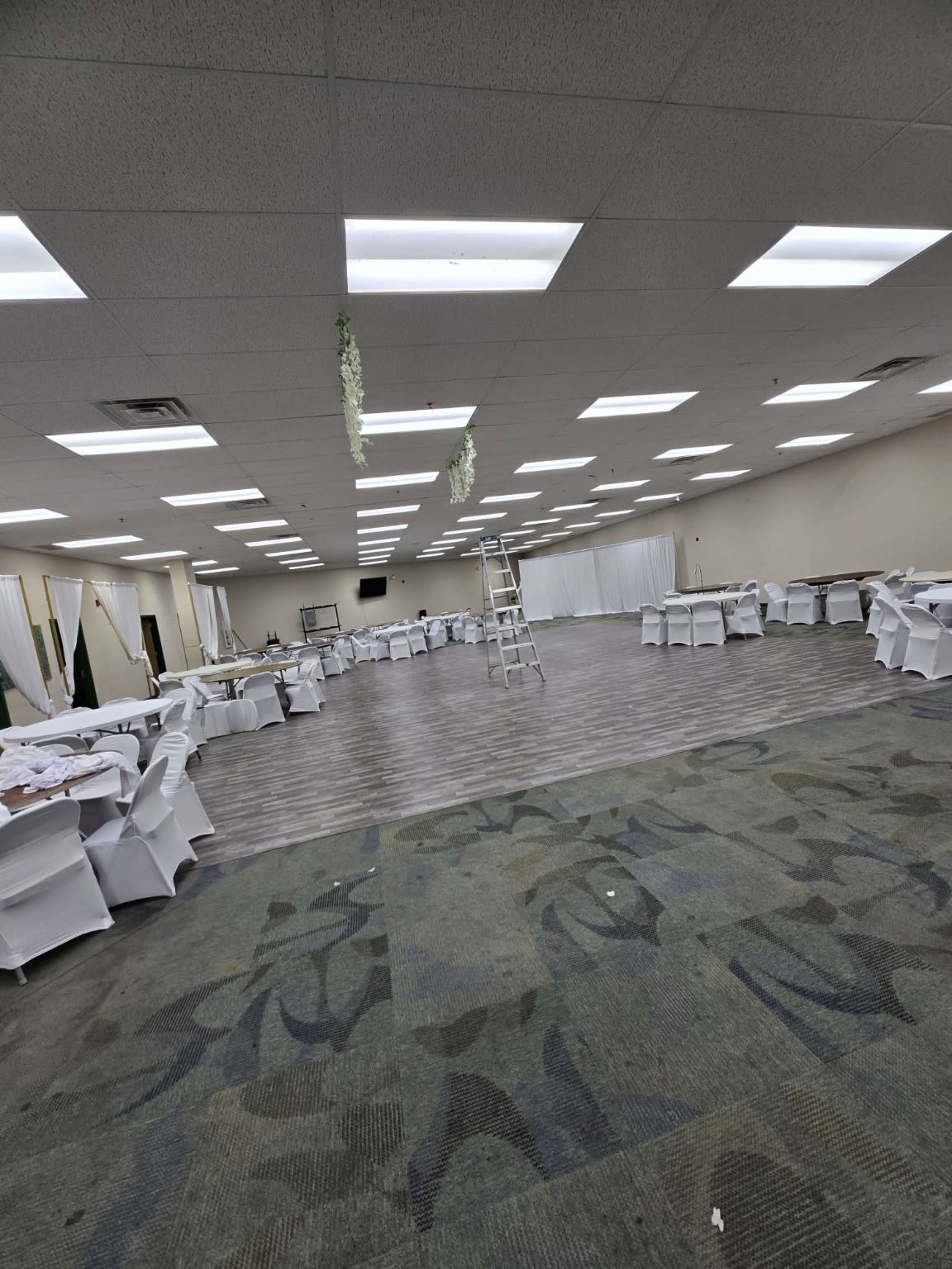 The image shows a large event space with multiple tables arranged in rows, a ladder positioned against a wall, and bright overhead lighting.