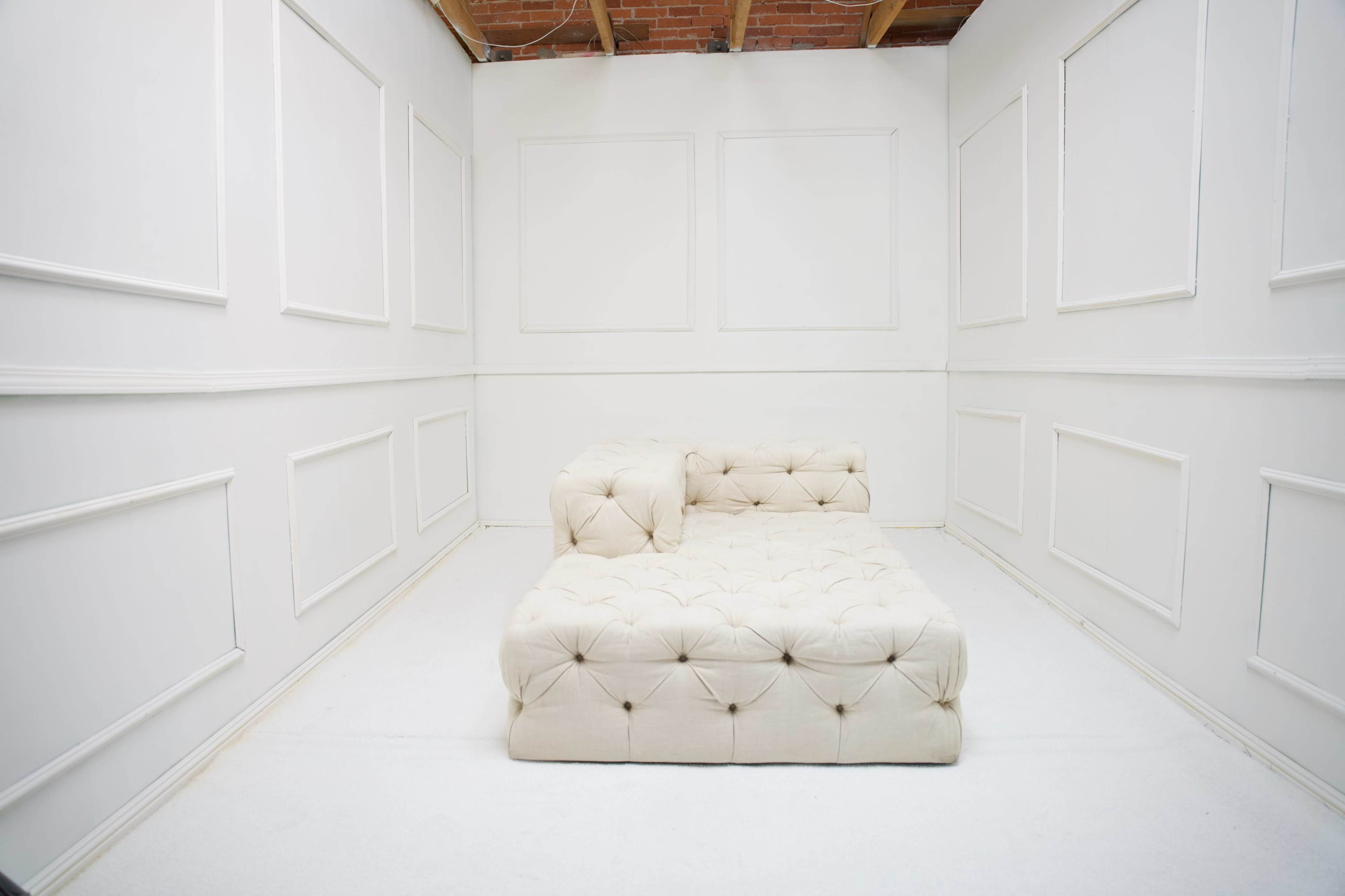 A light-colored tufted sofa is positioned in a sparsely decorated white room with plain walls and a ceiling featuring exposed beams.
