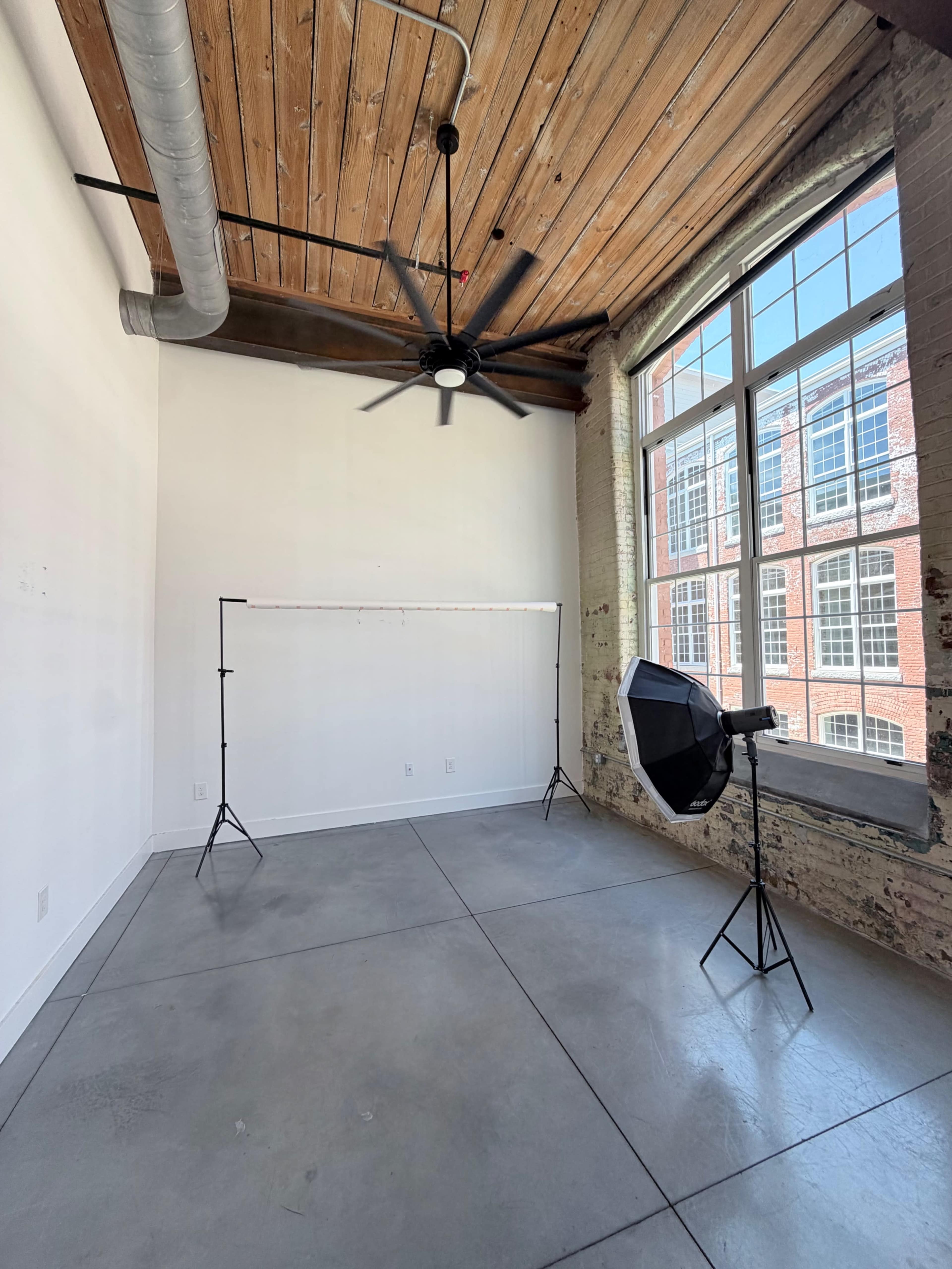 The image shows an empty room with a high ceiling, exposed wooden beams, a large window, a fan, a backdrop stand, and a light source setup.