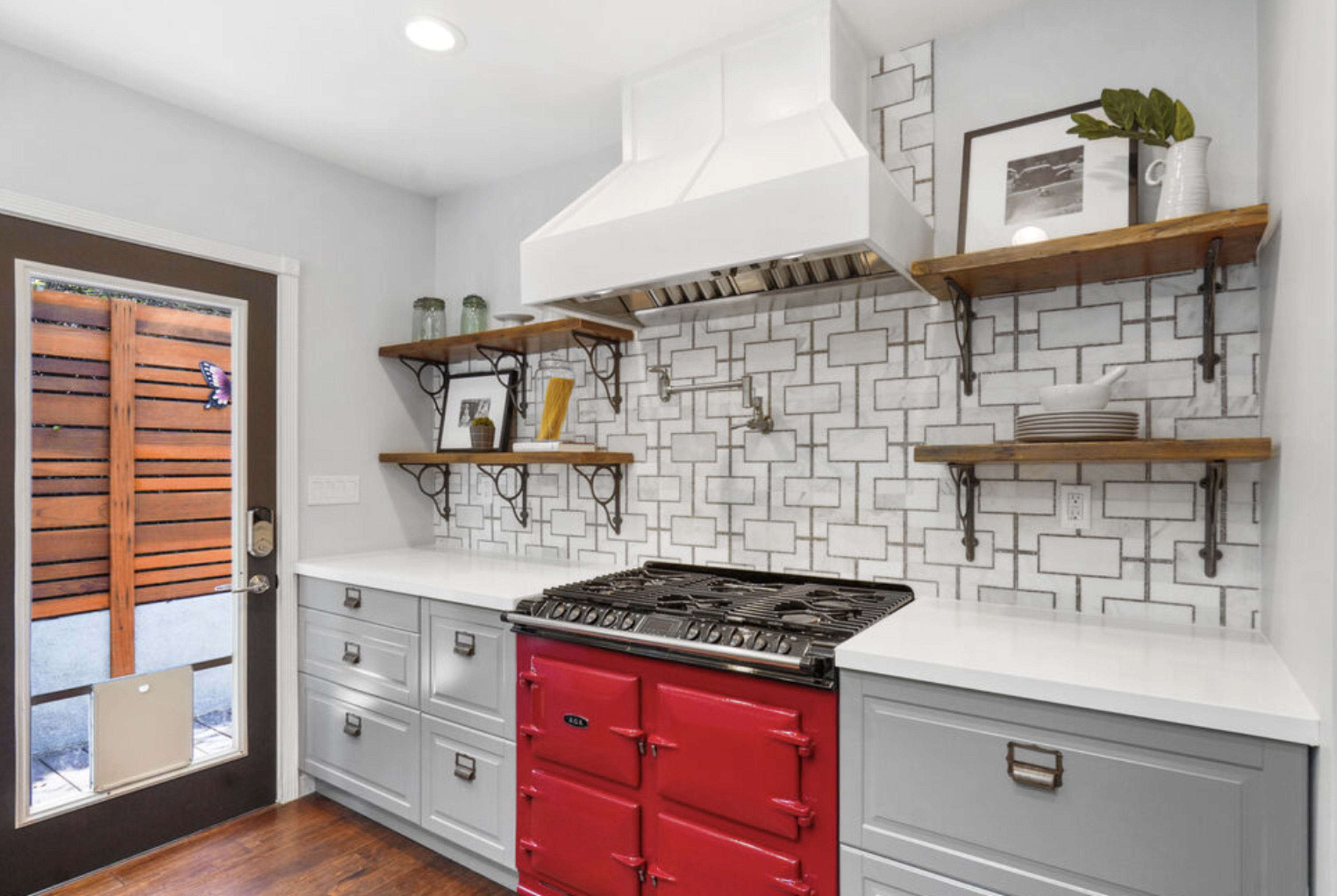A modern kitchen features a red gas range, white countertops, and open shelves against a geometric tile backsplash.