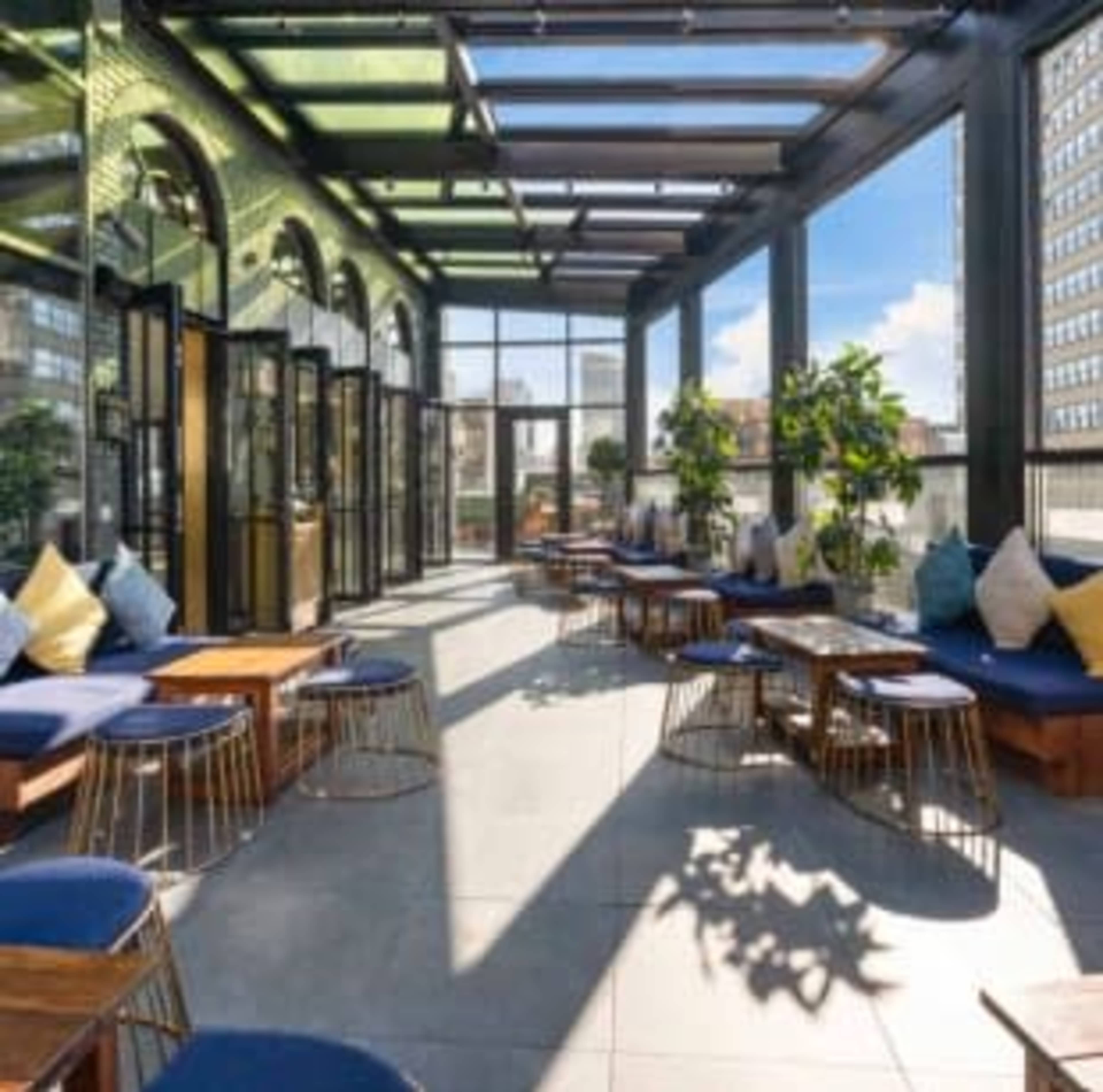 The image shows a brightly lit glass-enclosed dining area with wooden tables, blue seating, and plants, overlooking a cityscape.