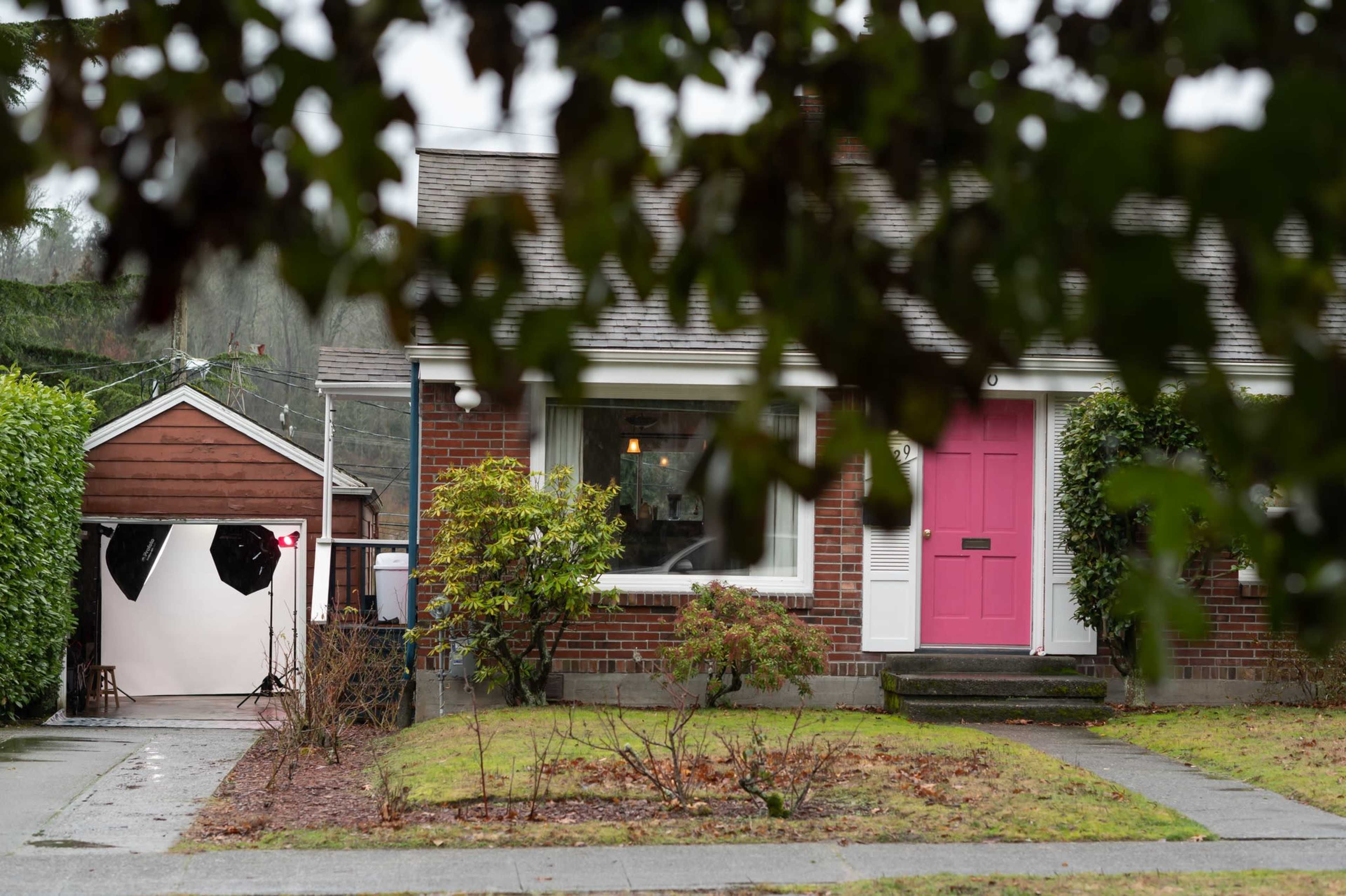 A brick house with a pink front door and a garage is partially obscured by foliage in a rainy setting.