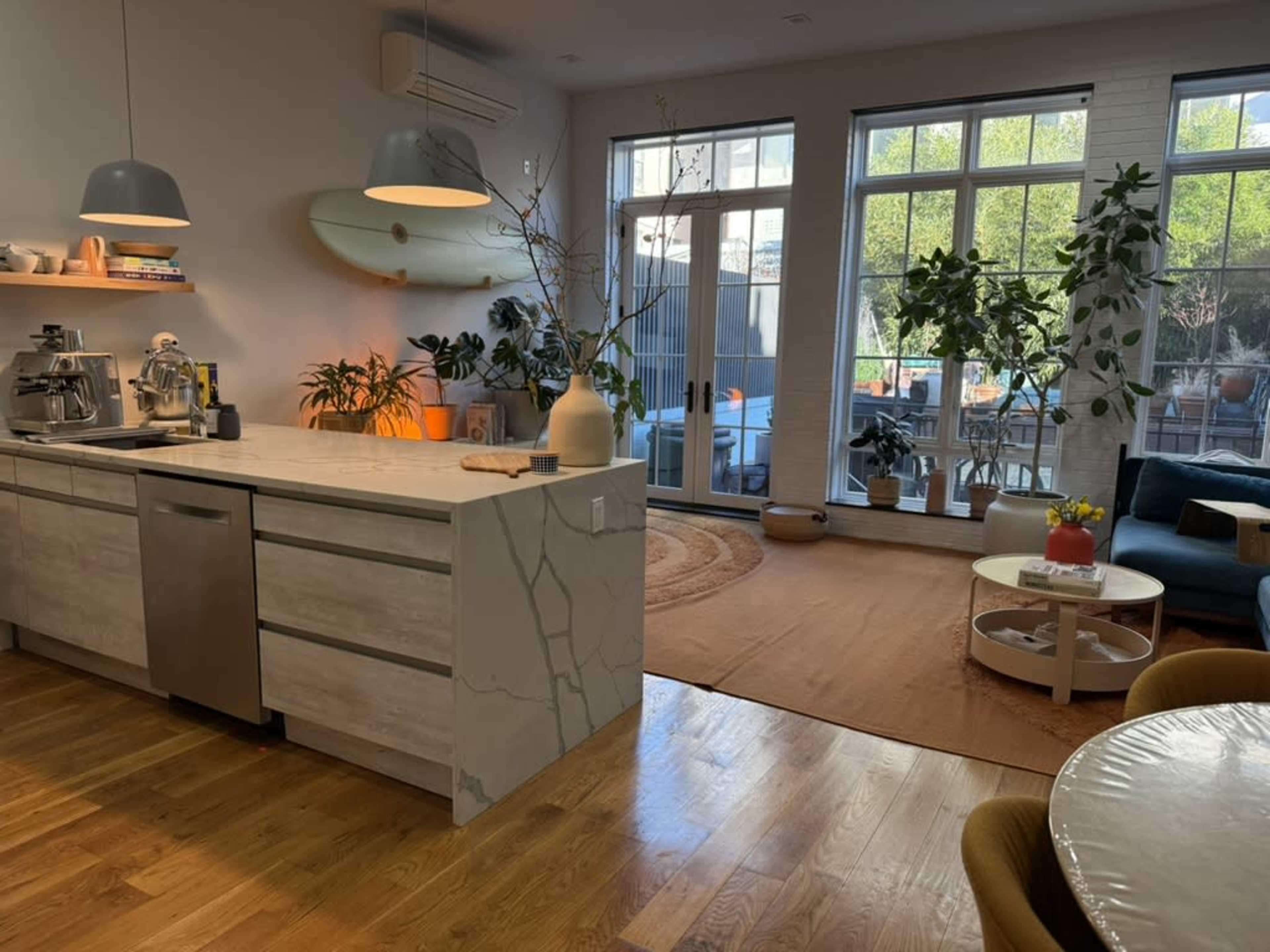 The image shows a modern kitchen and living area with large windows, plants, and a cozy seating arrangement.