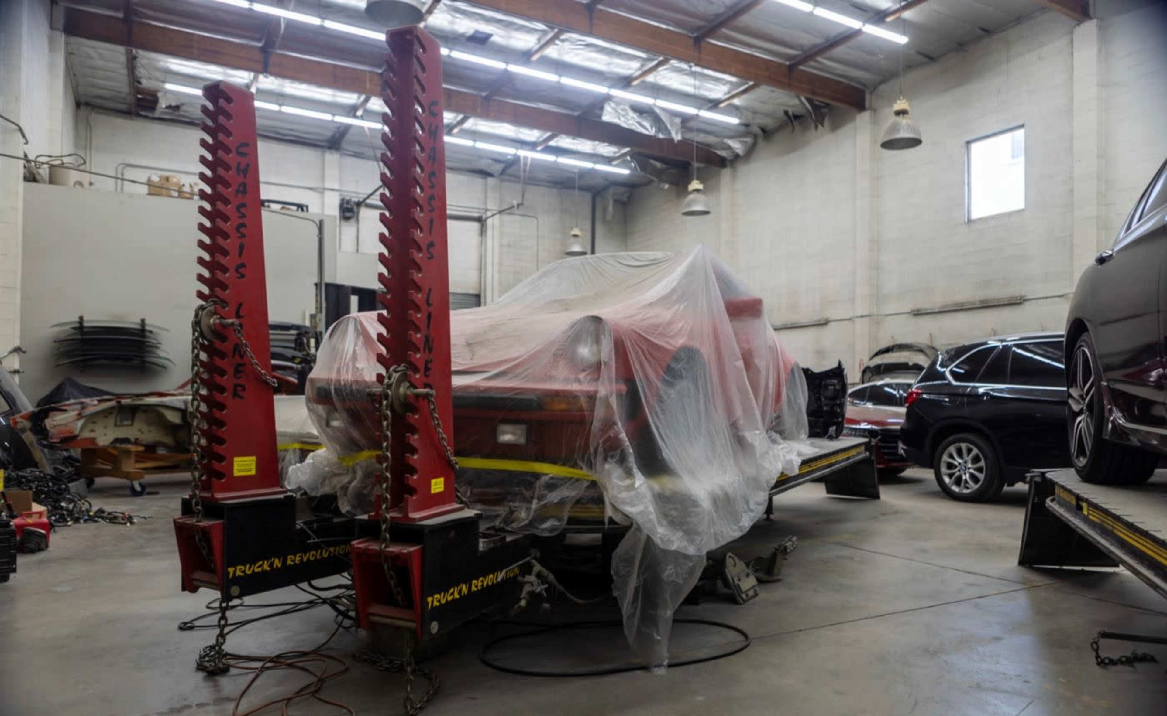 A red car is covered with a plastic sheet and mounted on a vehicle lift inside an auto repair shop.