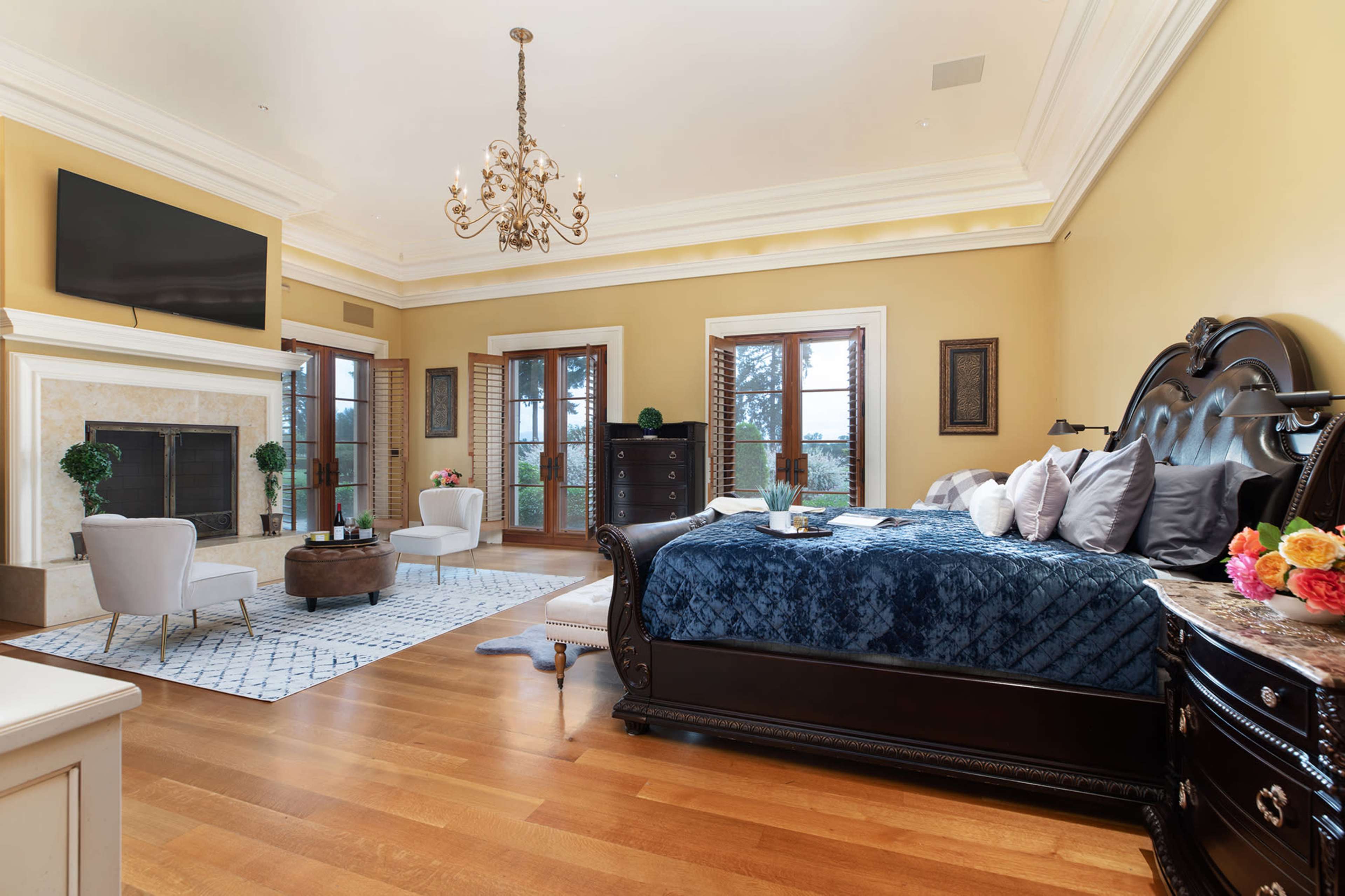 A spacious bedroom features a large bed, a chandelier, a fireplace, and a sitting area with two chairs and a decorative rug.