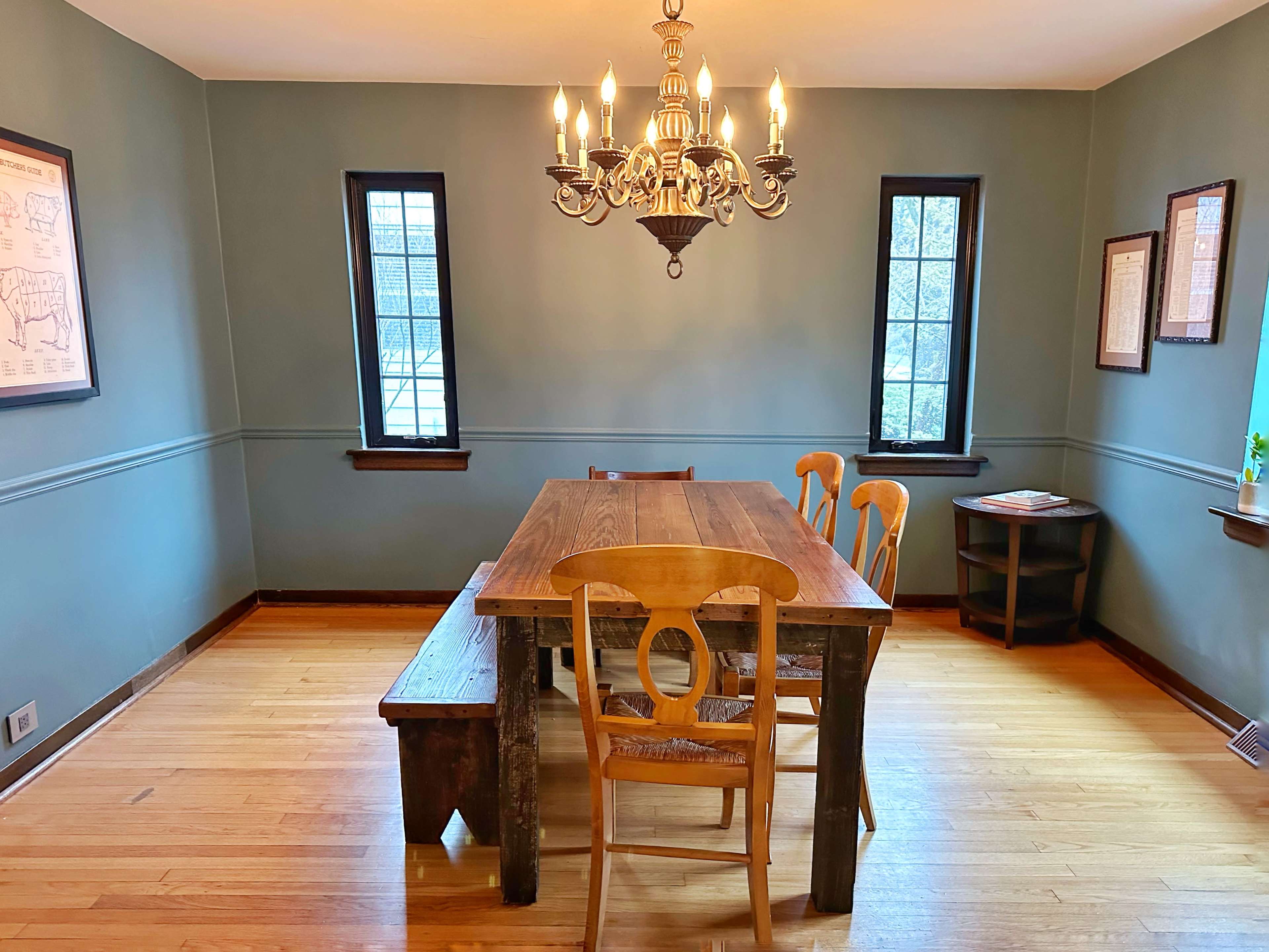 A wooden dining table is set in a room with two windows and a chandelier, surrounded by chairs and a bench.