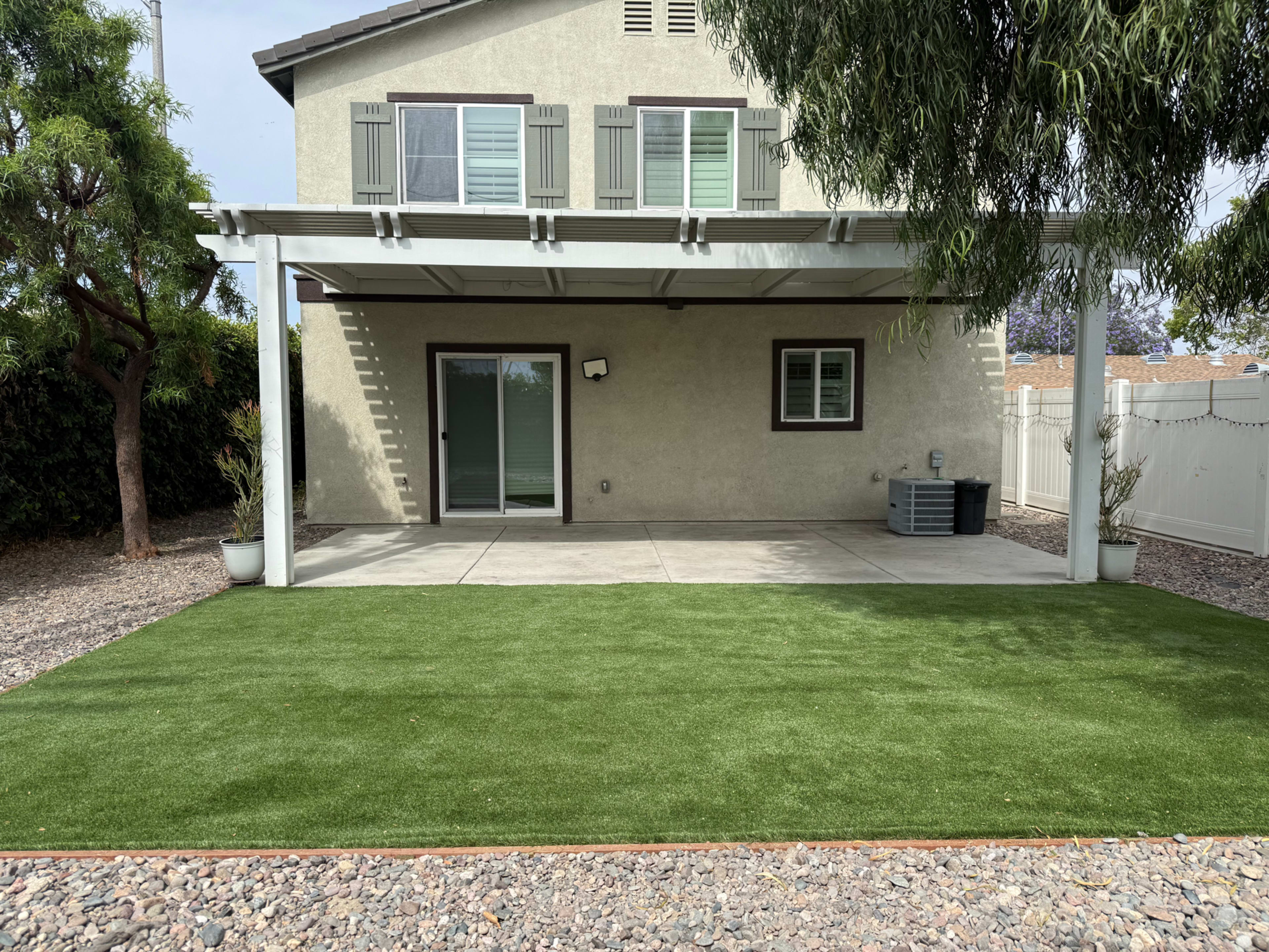 The image shows a house with a covered patio area and artificial grass in the foreground, surrounded by gravel and a fence.