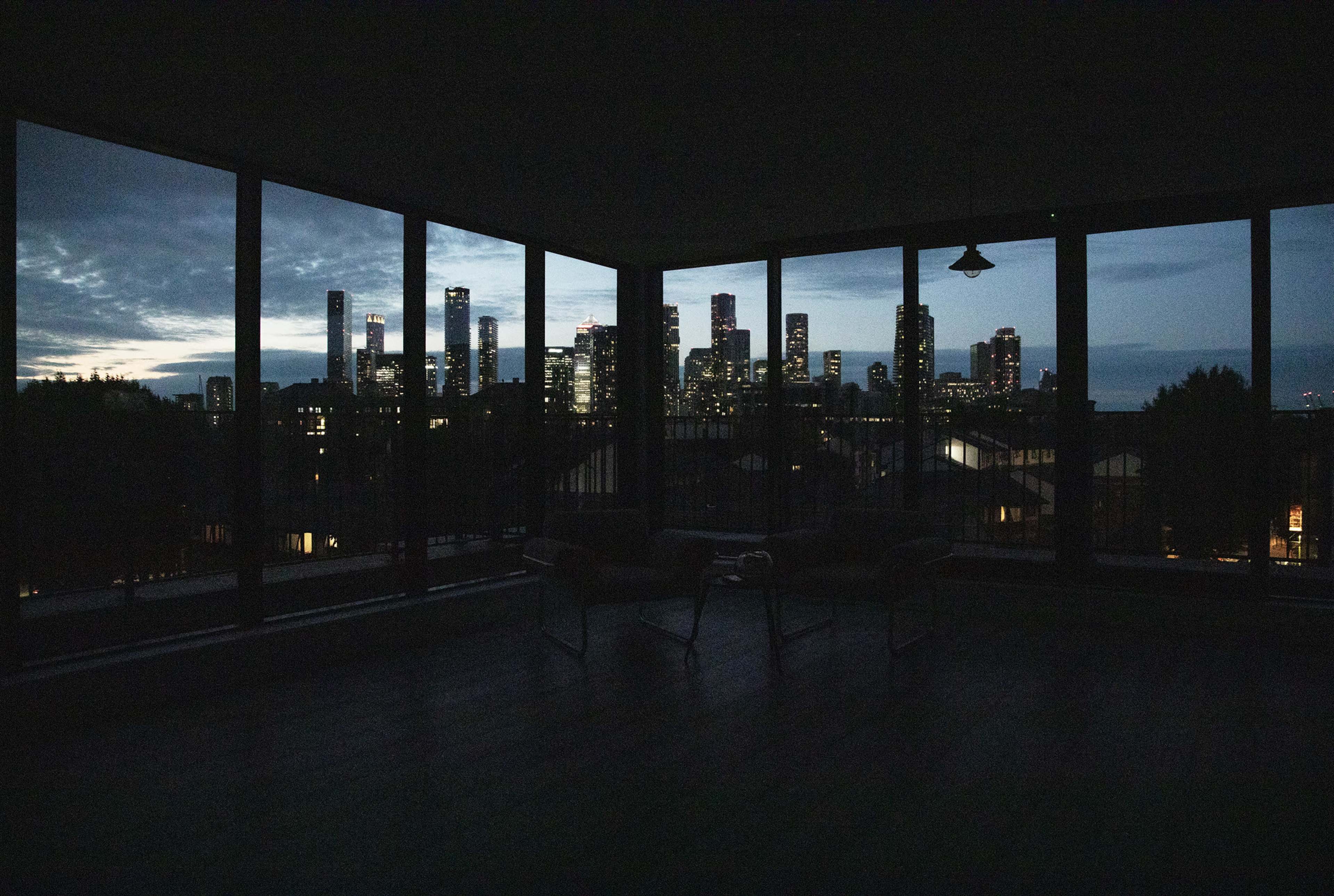A spacious room with large windows offers a view of a city skyline illuminated at dusk.