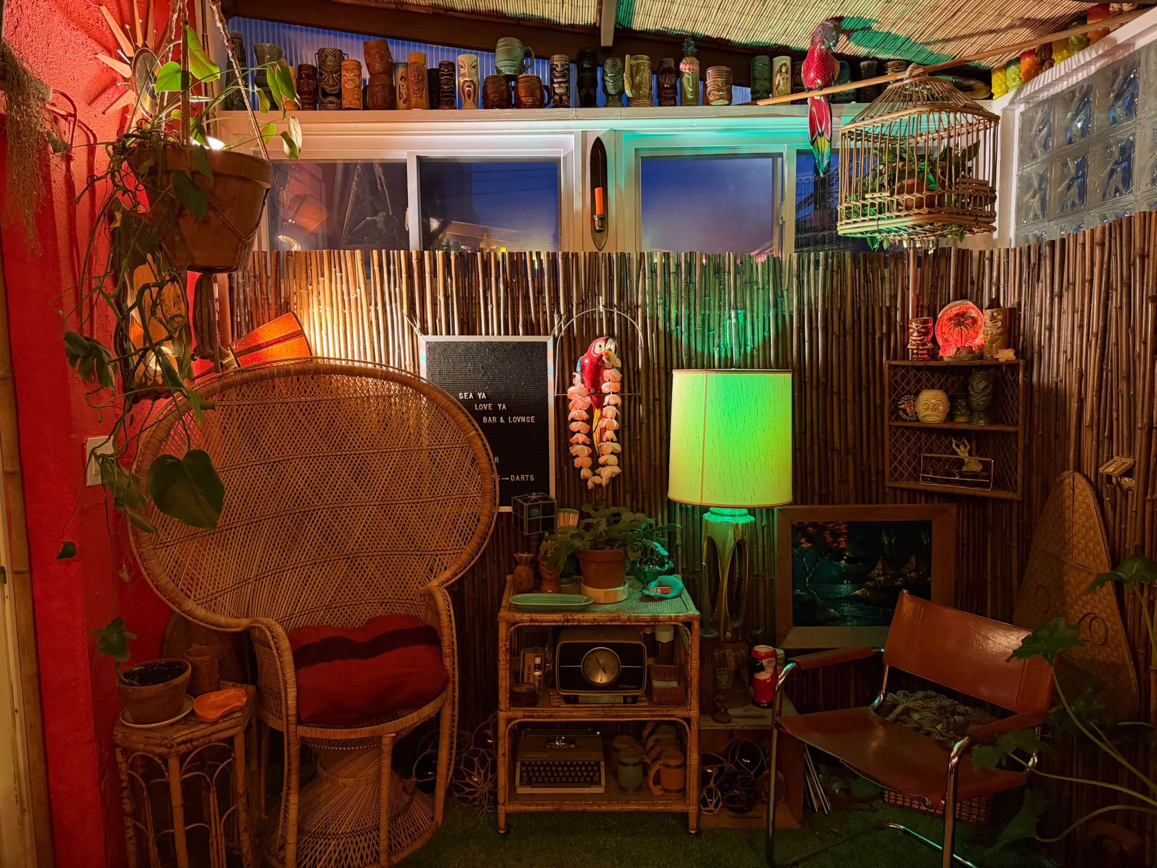 The image shows a cozy interior space decorated with plants, colorful lighting, and vintage furniture including a large rattan chair and a wooden chair.