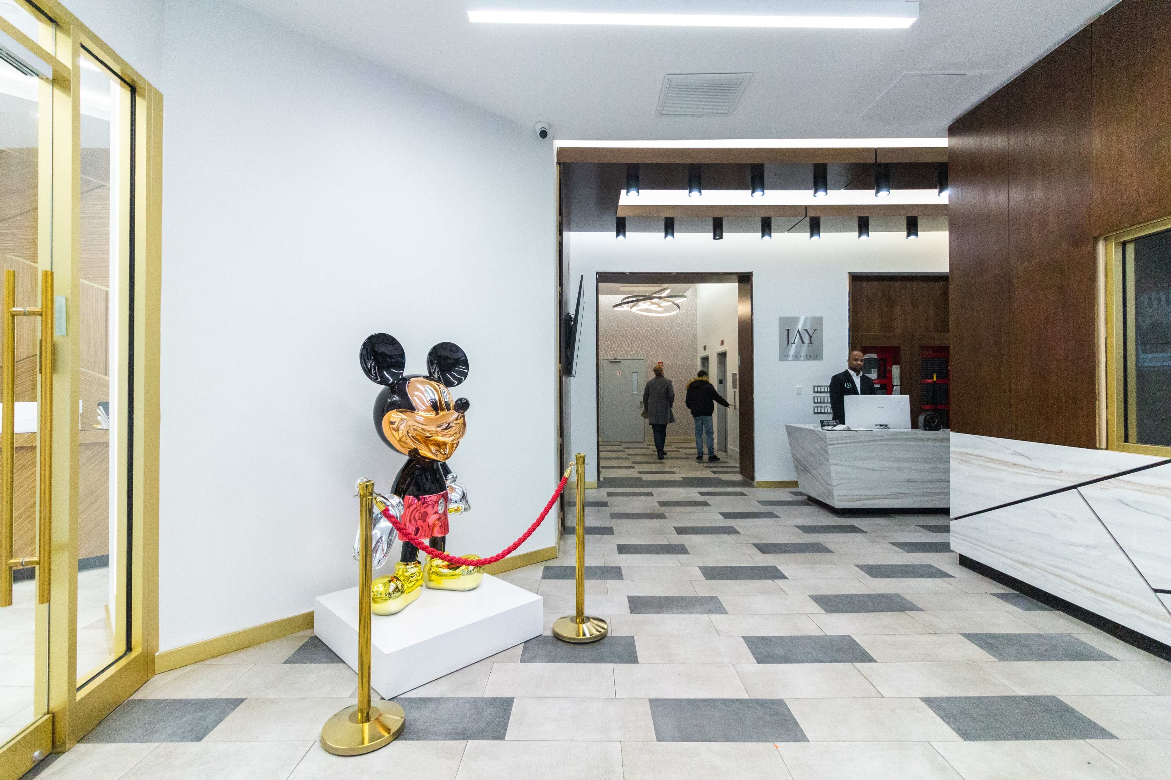 The image shows a modern lobby featuring a decorative Mickey Mouse sculpture on a pedestal, flanked by a reception desk and visitors walking in the background.