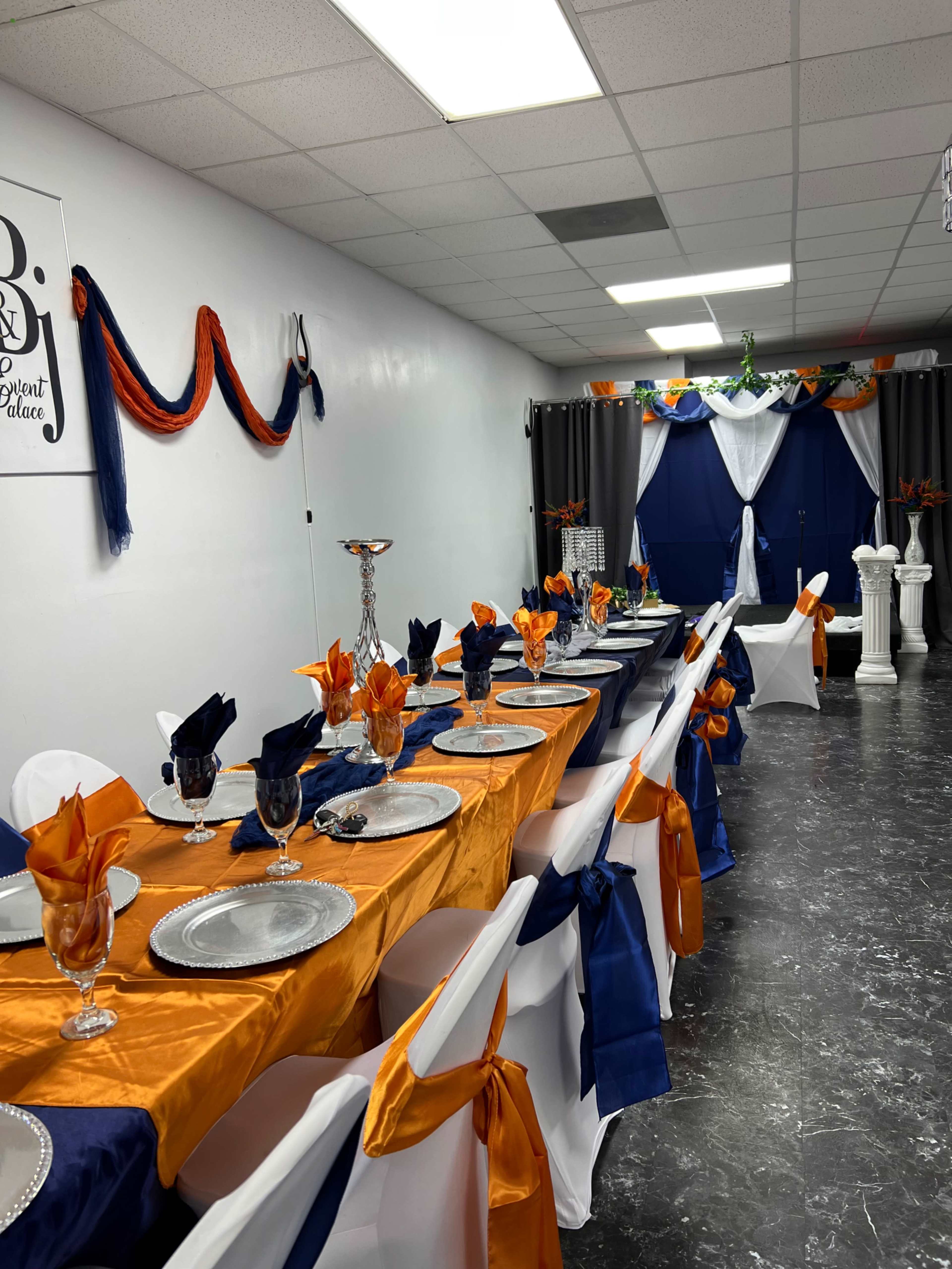 A decorated event space features a long table dressed in orange and navy fabric, with chairs adorned in white covers and colorful sashes, set for a gathering.