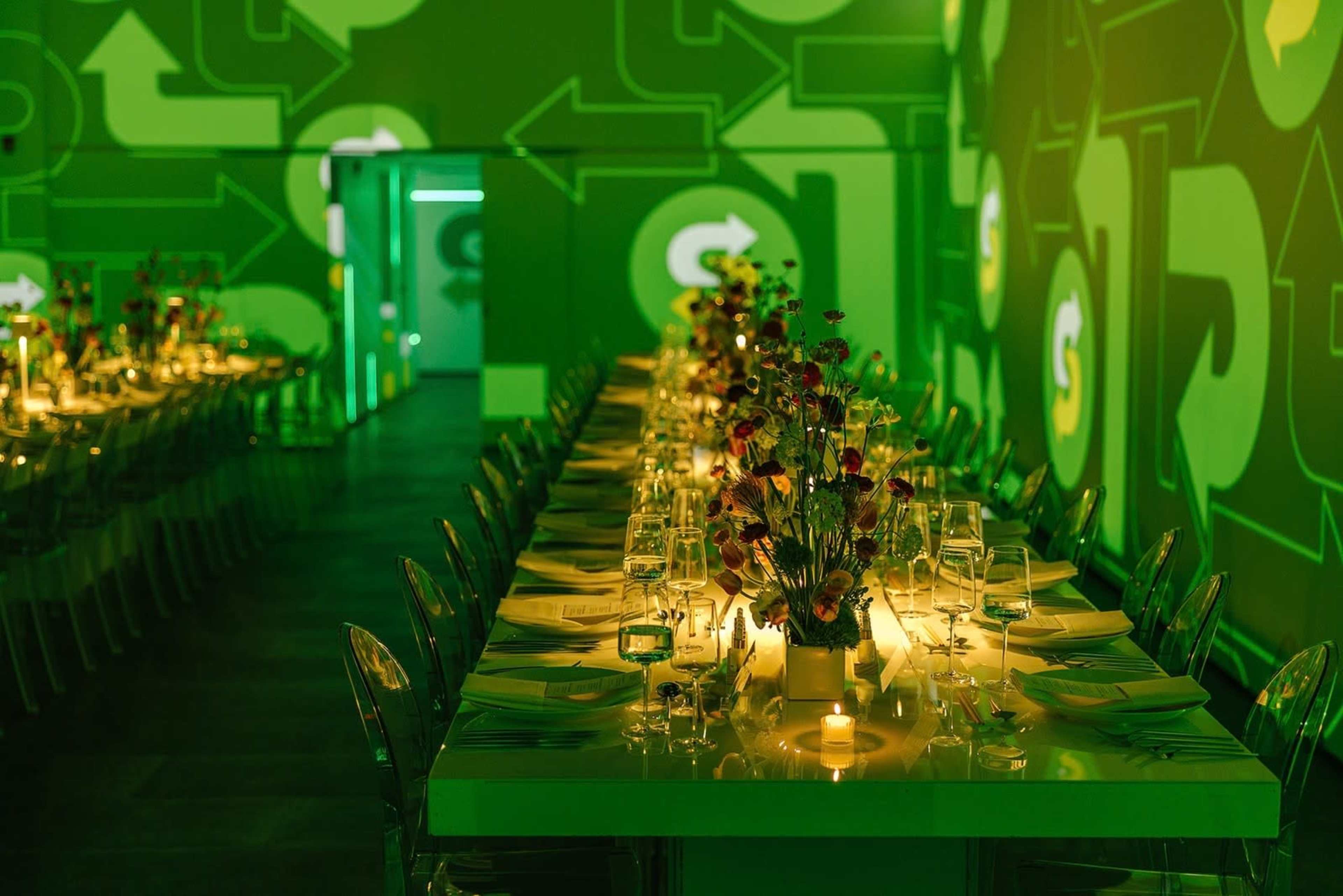 A long, elegantly set dining table is lit by glowing candles and adorned with flowers, against a vibrant green wall featuring abstract designs.