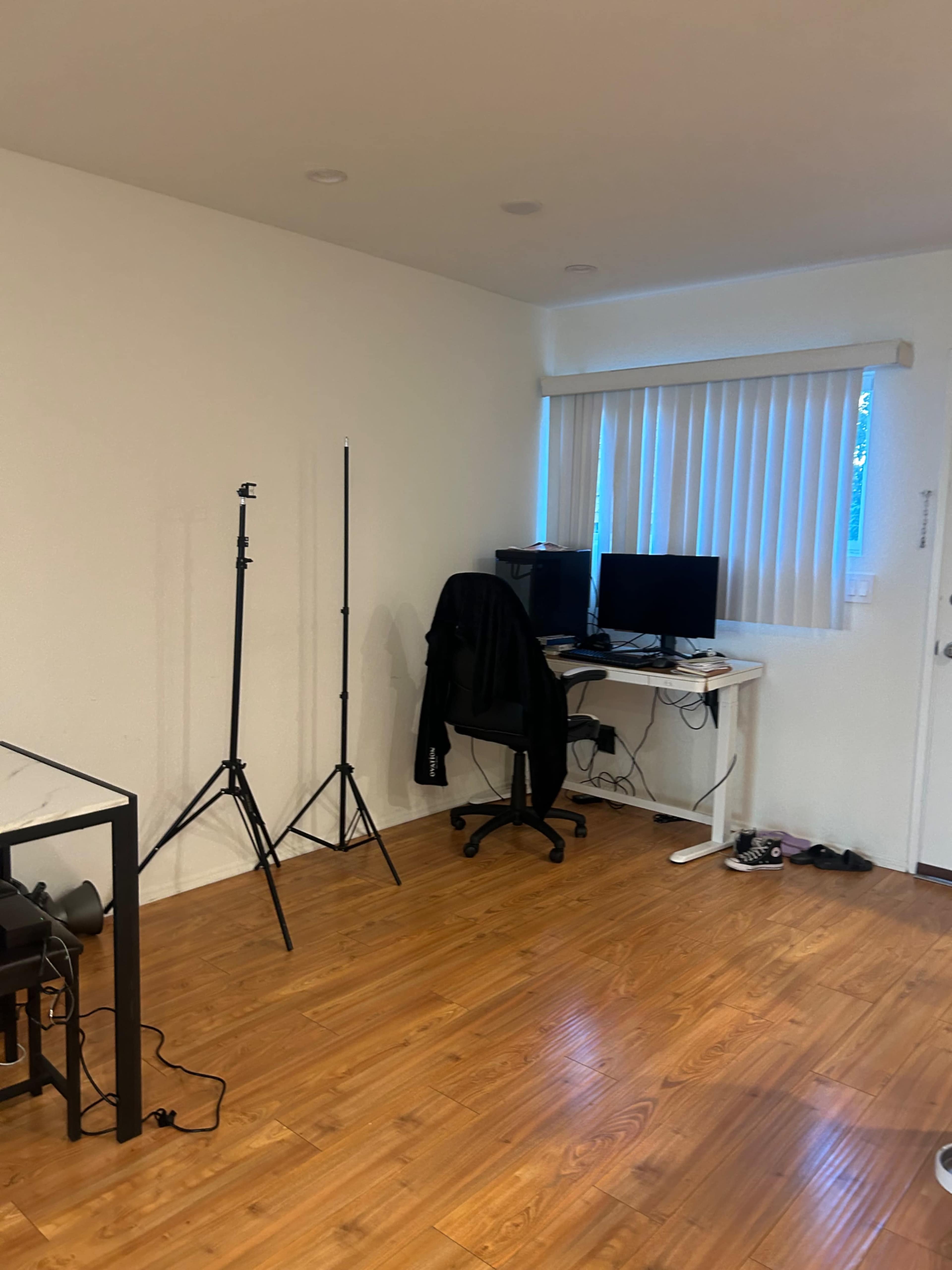 The image shows a sparsely furnished room with wooden flooring, featuring a desk with two monitors, a black chair, and a couple of stands, along with a few items casually placed on the floor.