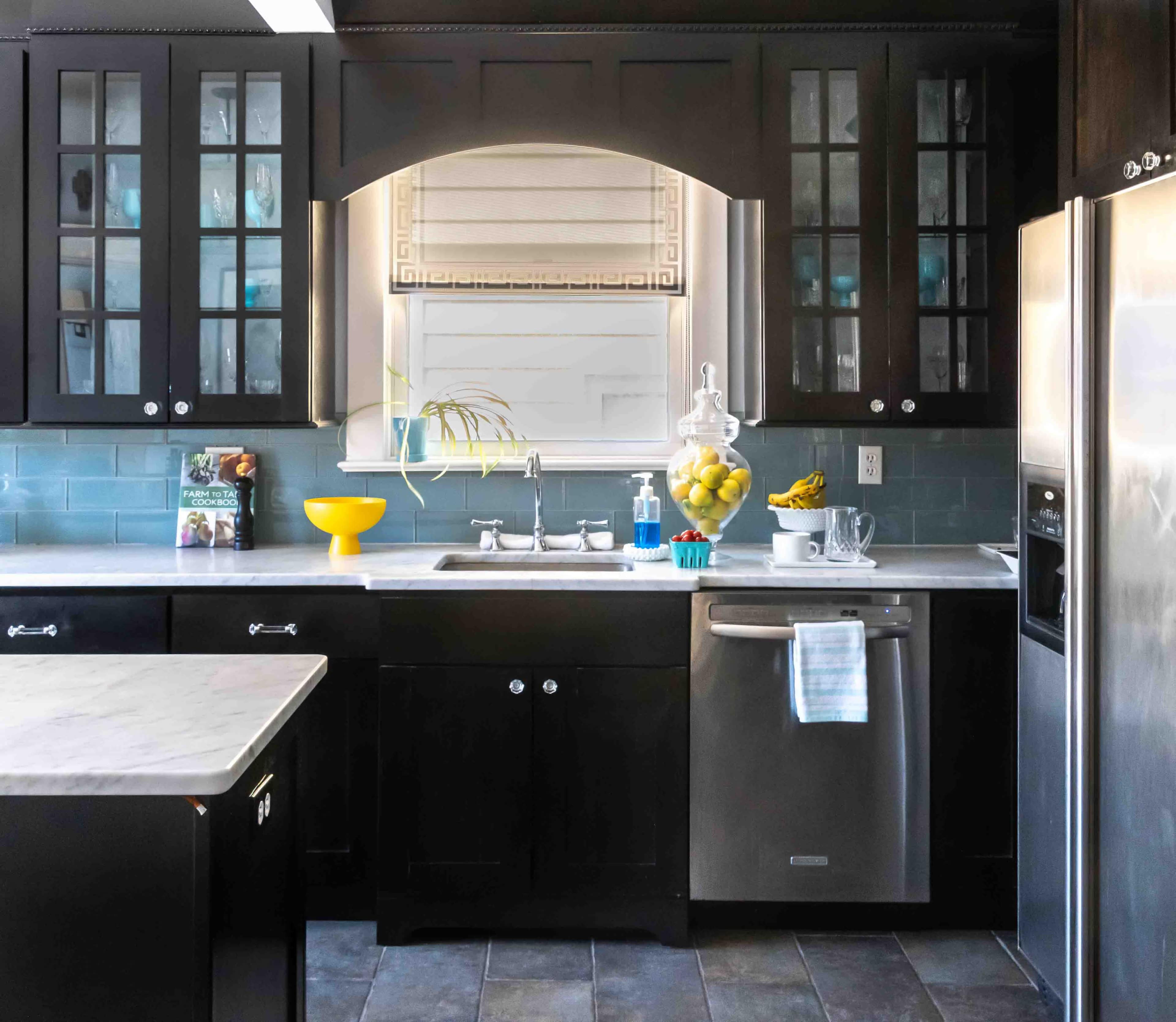 The image shows a modern kitchen with dark cabinetry, a stainless steel refrigerator, and a marble countertop, featuring a window above the sink and decorative items on display.