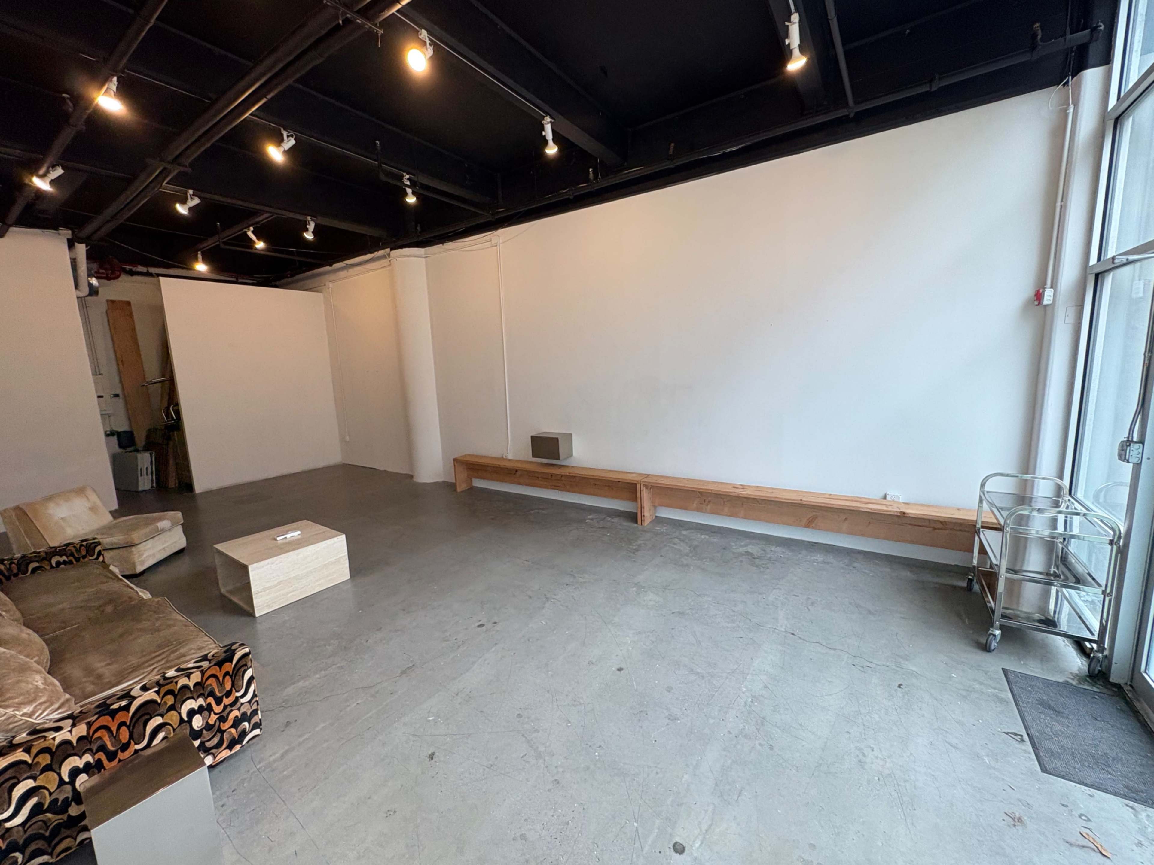 The image shows a spacious, empty room with a concrete floor, a wooden bench along one wall, a small table in the center, and a seated area with a couch.