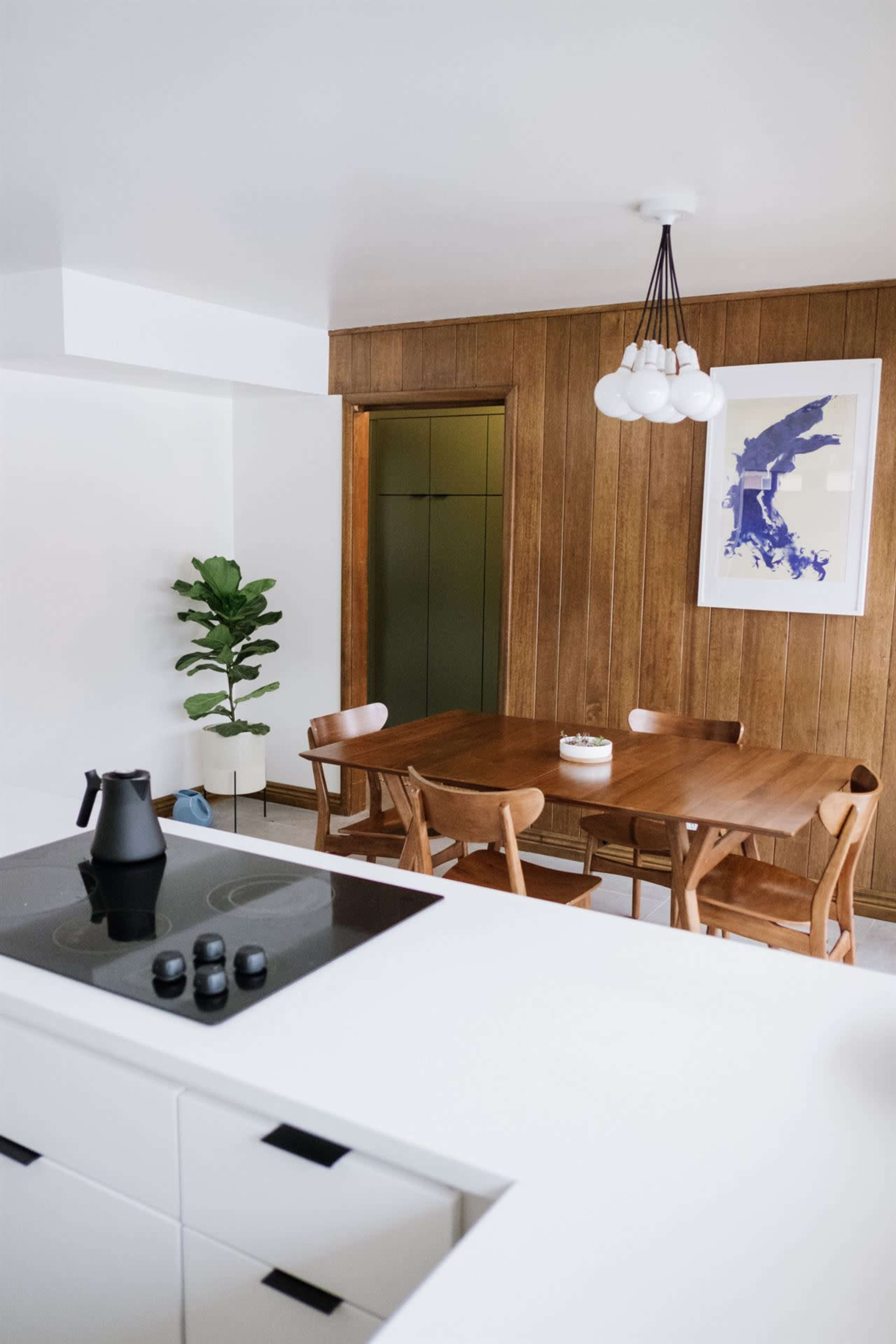A modern kitchen and dining area featuring a wooden table with chairs, a potted plant, and contemporary light fixtures.