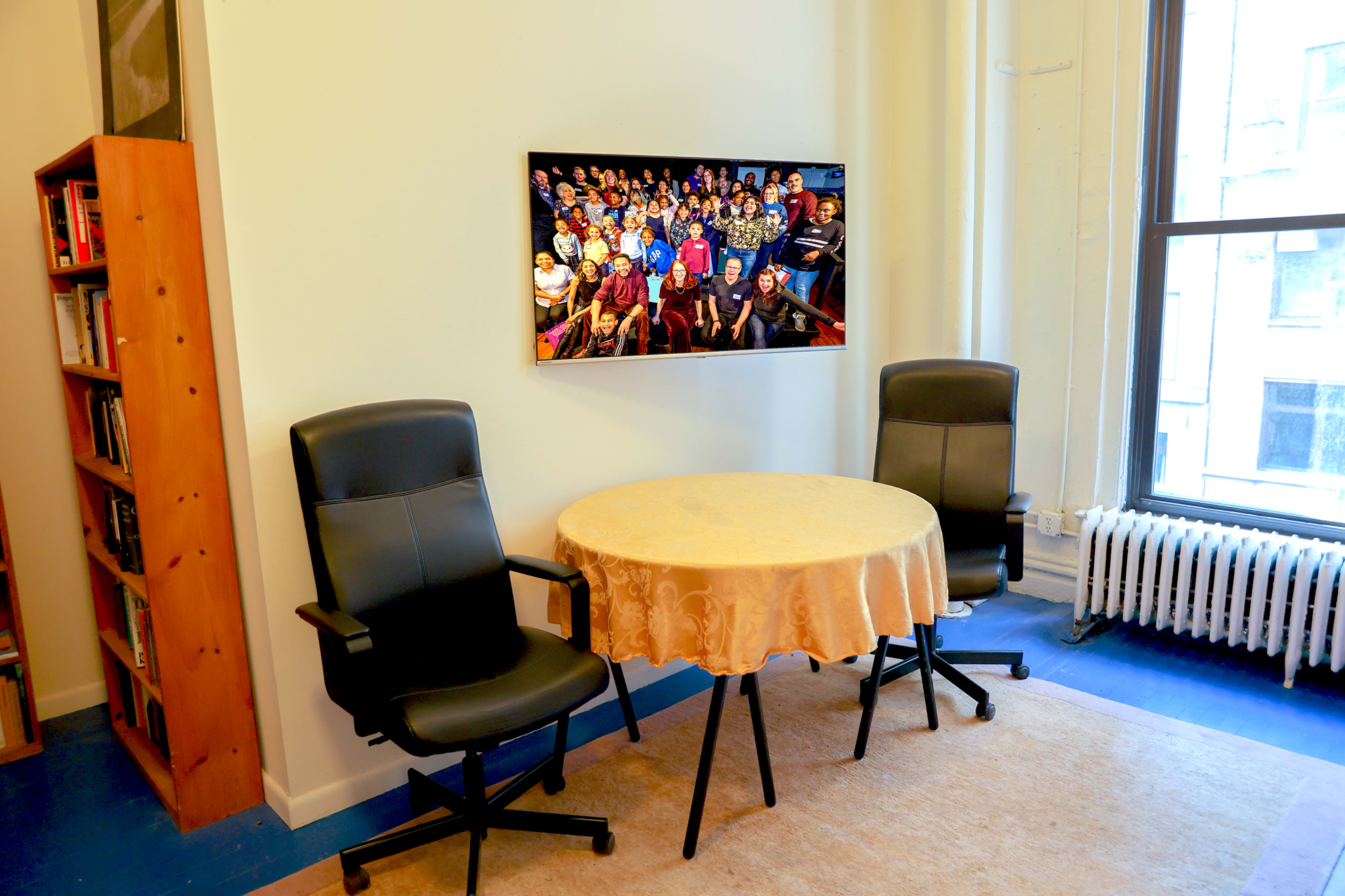 A small, brightly lit room features a round table with a tablecloth and two black chairs, alongside a wall-mounted television displaying a group photo.